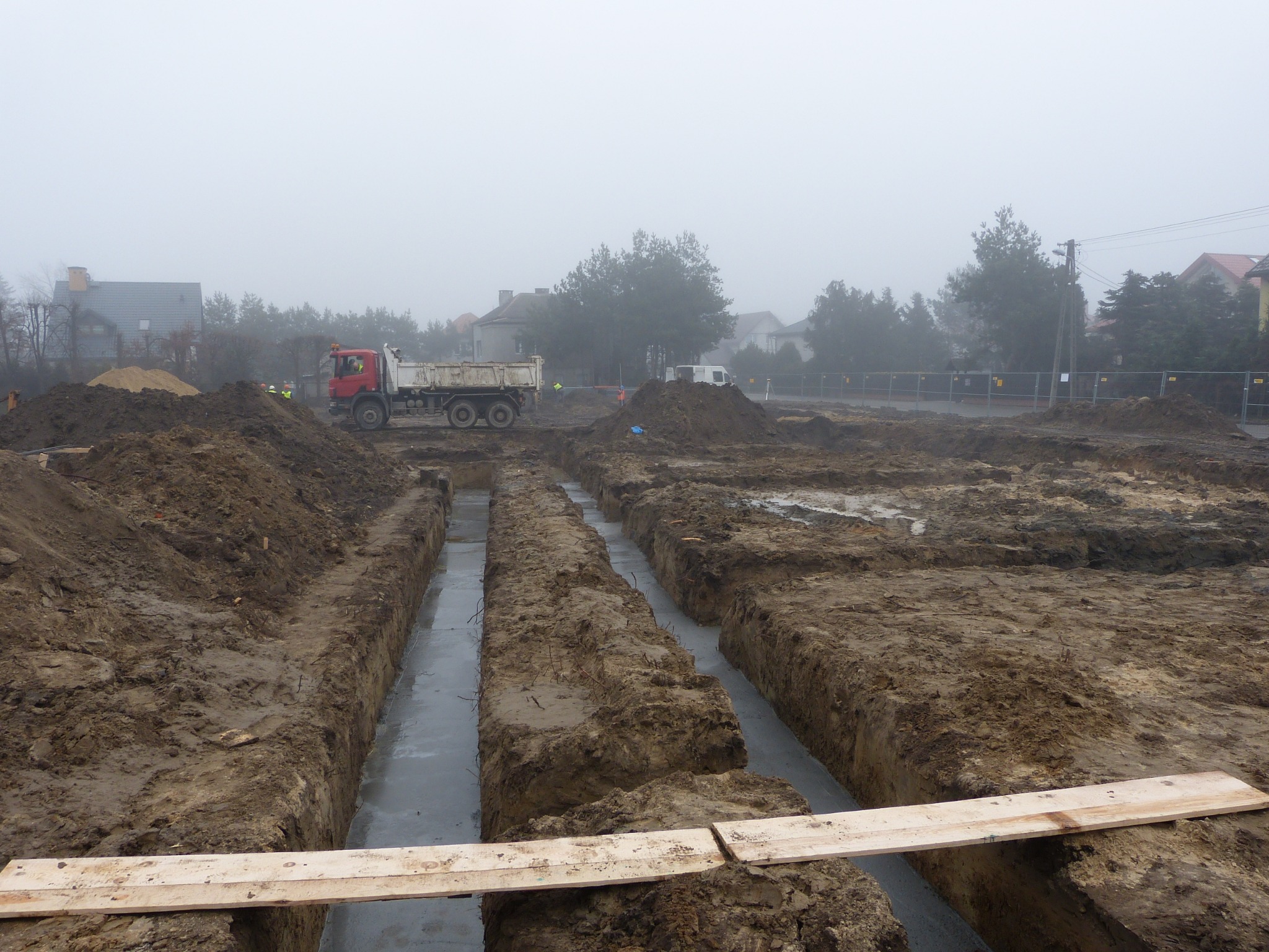 Wykopane rowy fundamentowe wypełnione betonem na placu budowy w mglisty dzień, widoczna ciężarówka i fragmenty zabudowy w tle.