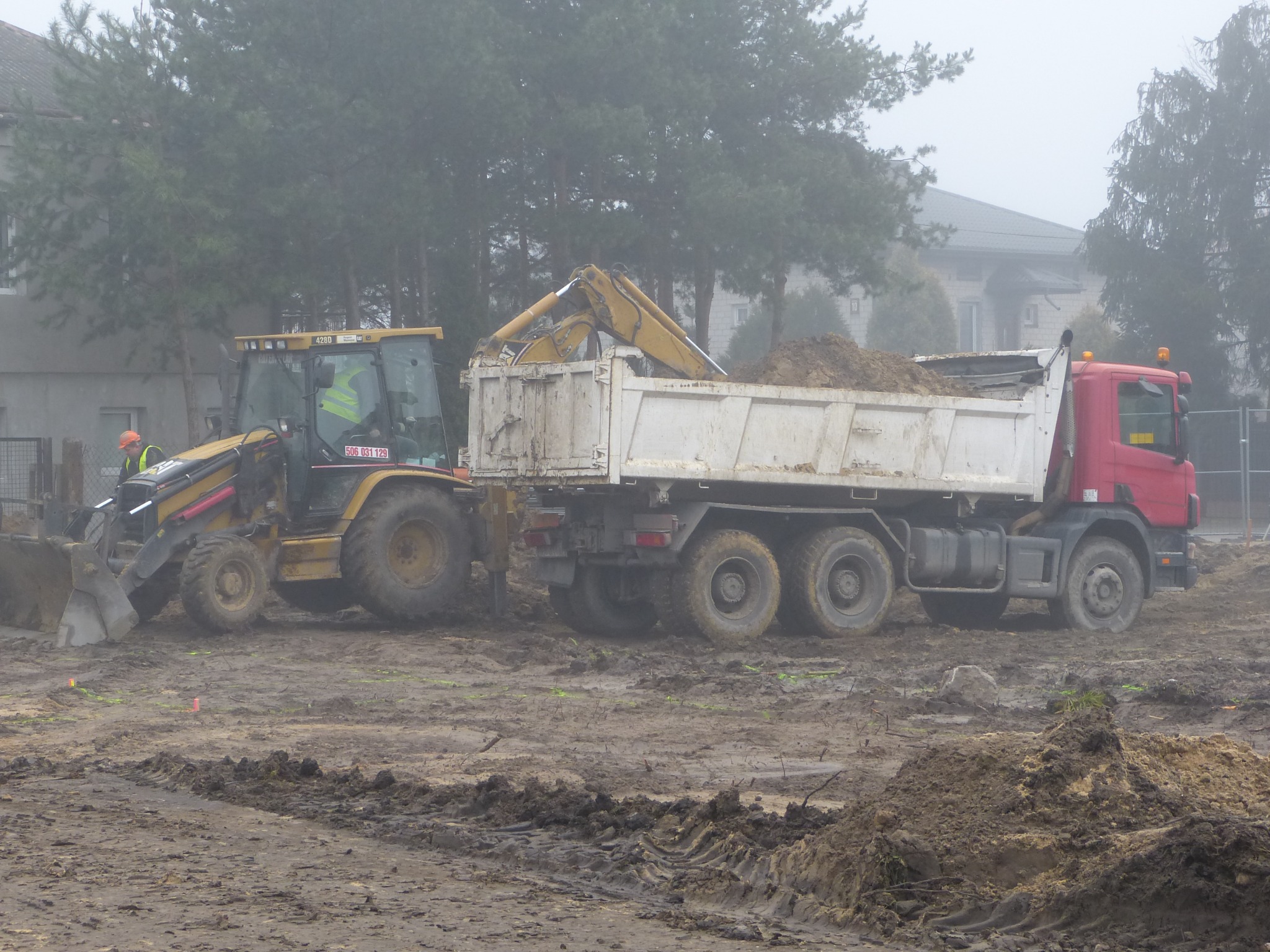 Żółta koparko-ładowarka nabiera ziemię do białej ciężarówki z czerwoną kabiną na rozmokłym placu budowy w mglisty dzień.