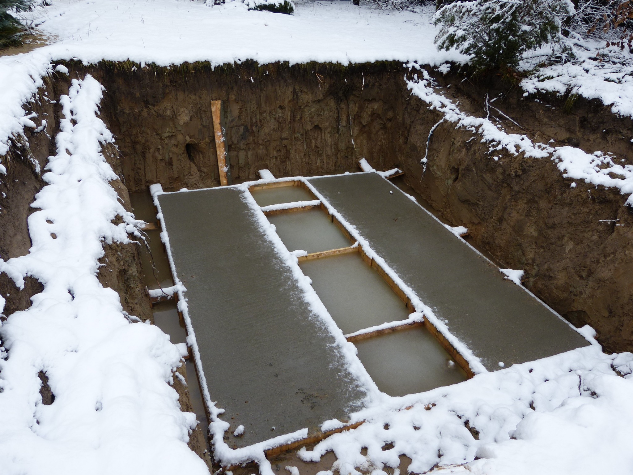 Świeżo wylany beton w wykopie, z drewnianym szalunkiem i śniegiem na krawędziach, zimowa sceneria.