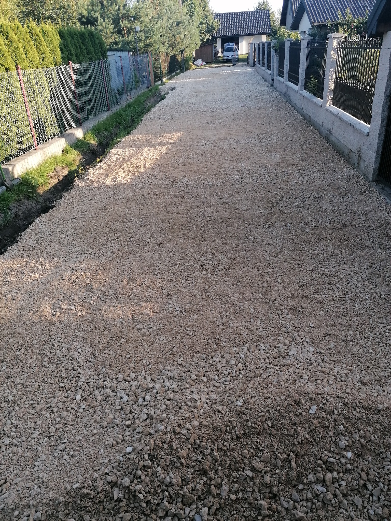 Świeżo wysypana, jasna droga szutrowa pomiędzy ogrodzeniami posesji, z widocznym rowem odwadniającym po lewej stronie i zaparkowanym samochodem w oddali.