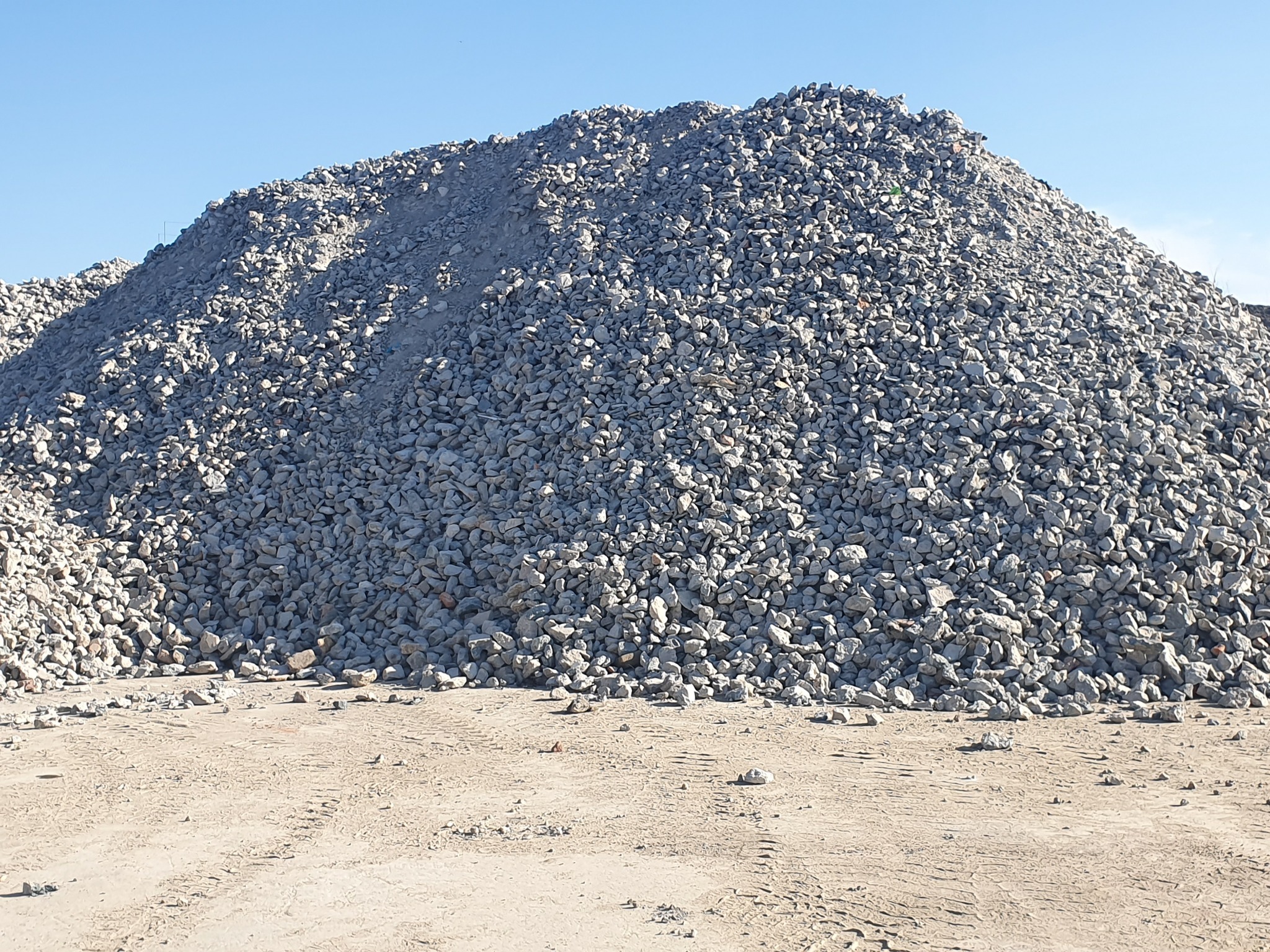 Góra szarego kruszywa na piaszczystym podłożu, widoczne ślady opon, błękitne niebo w tle.