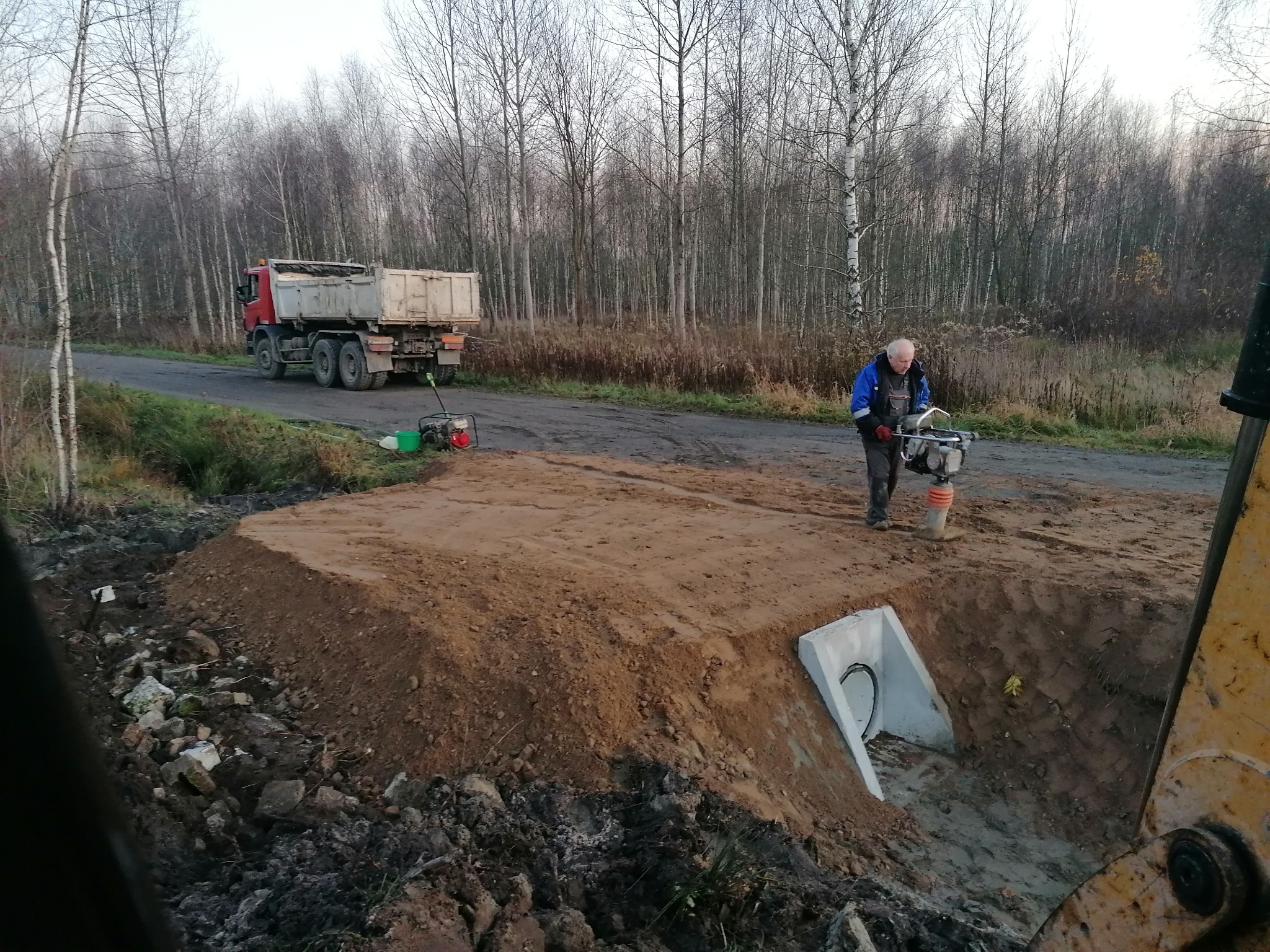Pracownik zagęszcza ubijarką wibracyjną nasyp ziemny nad betonowym przepustem, w tle wywrotka i rzadki las.