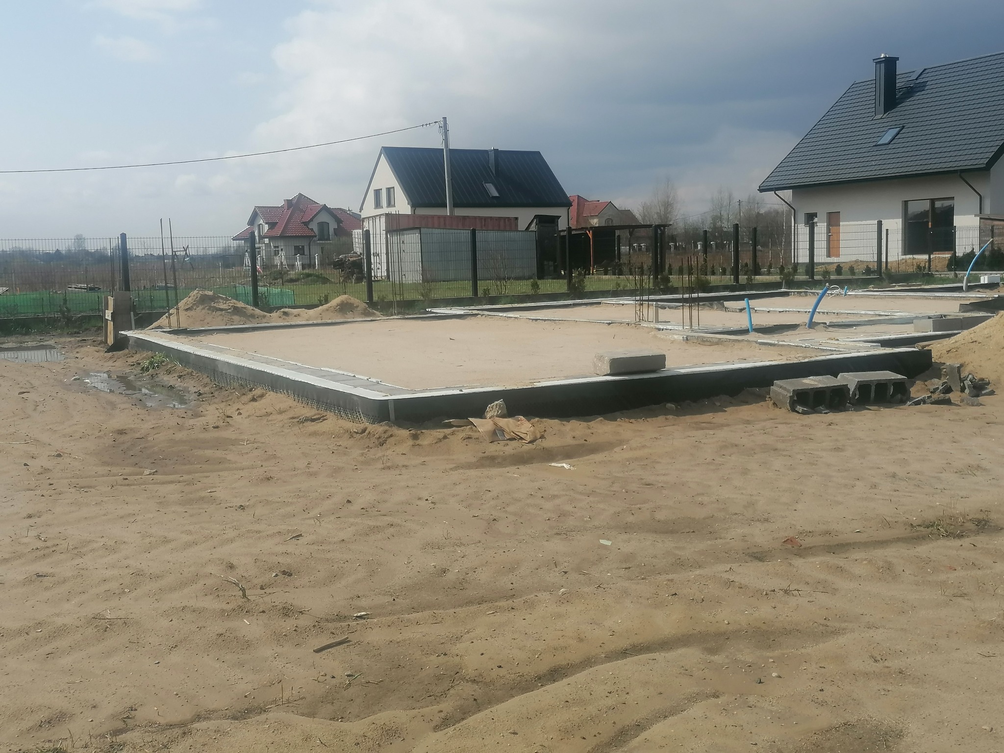 Przygotowany teren pod budowę domu jednorodzinnego, widoczne fundamenty i piasek, w tle zabudowa mieszkalna i pochmurne niebo.