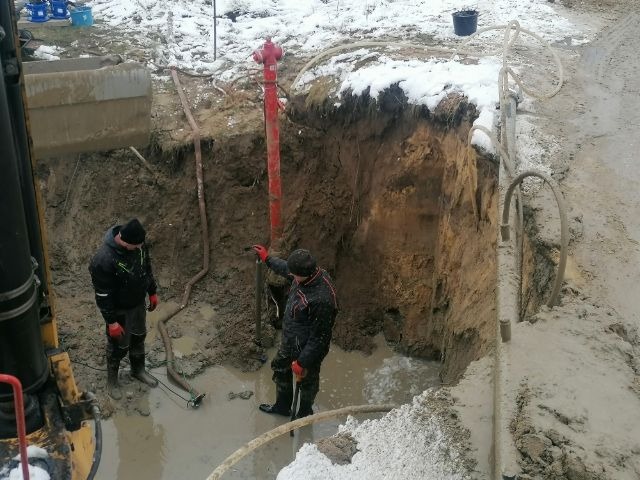 Dwóch pracowników w czarnych kurtkach i kaloszach stoi w wykopie wypełnionym wodą, obok czerwonego hydrantu i węży, na częściowo ośnieżonym terenie.