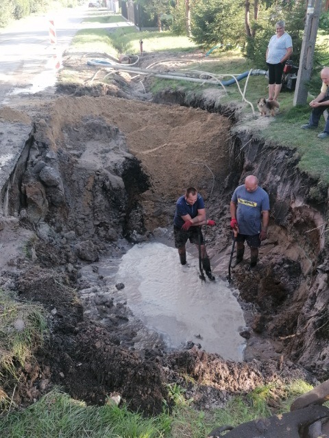 Głęboki wykop w ziemi wypełniony wodą, dwóch mężczyzn w gumowcach stoi wewnątrz, trzymając łopaty, na powierzchni widoczny uszkodzony asfalt, w tle ulica i obserwujący ludzie z psem.
