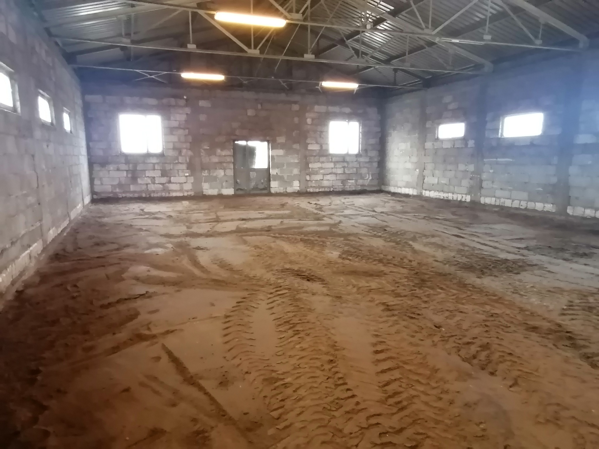Wnętrze hali z betonowych bloków, ziemia na podłodze z widocznymi śladami opon, stalowa konstrukcja dachu i rzędy świetlówek.