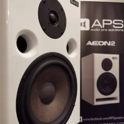 Biały głośnik studyjny APS Aeon2 z czarnymi elementami, widoczny na tle baneru reklamowego APS Audio Pro Solutions.