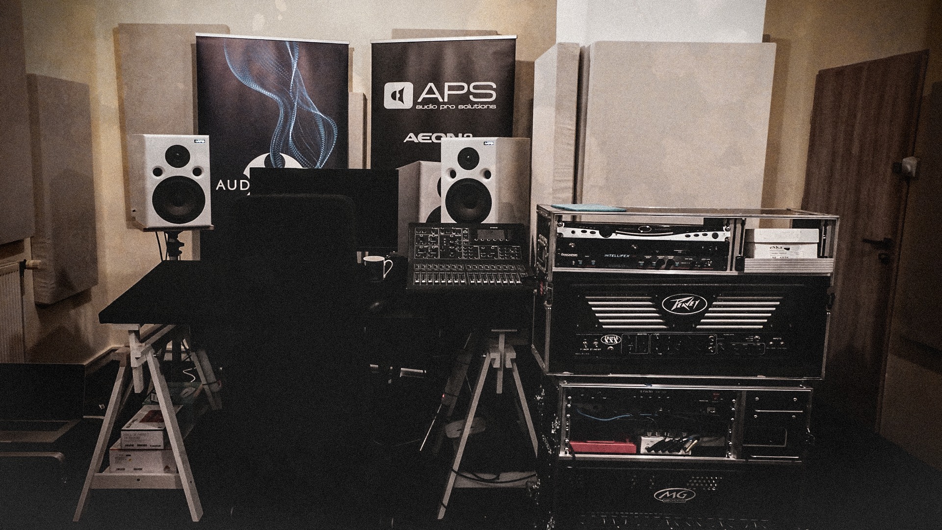 Panel mikserski na biurku w pomieszczeniu z panelami akustycznymi i kolumnami głośnikowymi, widoczne również urządzenia rackowe i banery reklamowe APS.