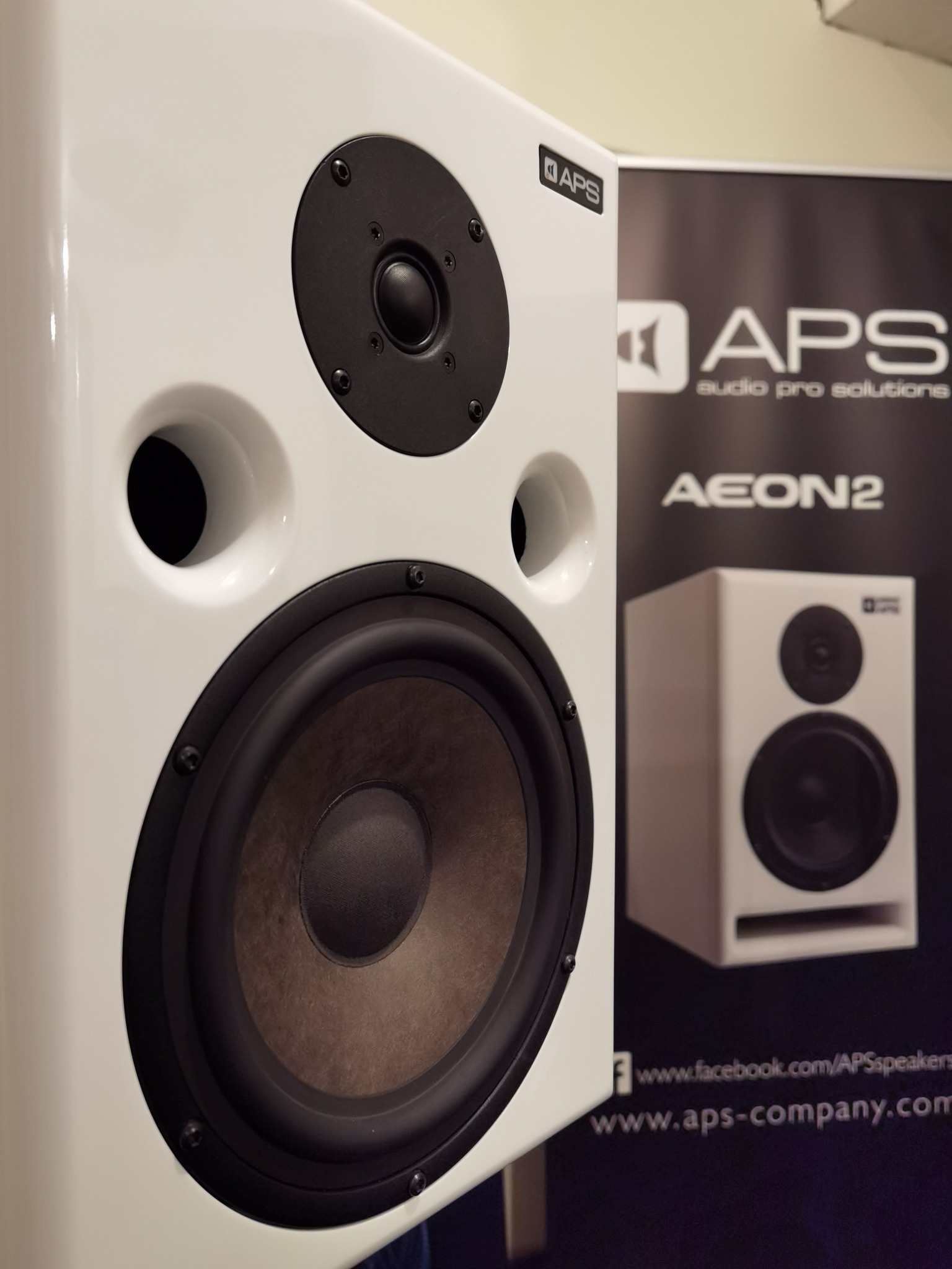 Biały głośnik studyjny APS Aeon2 z czarnymi elementami, widoczny na tle baneru reklamowego APS Audio Pro Solutions.