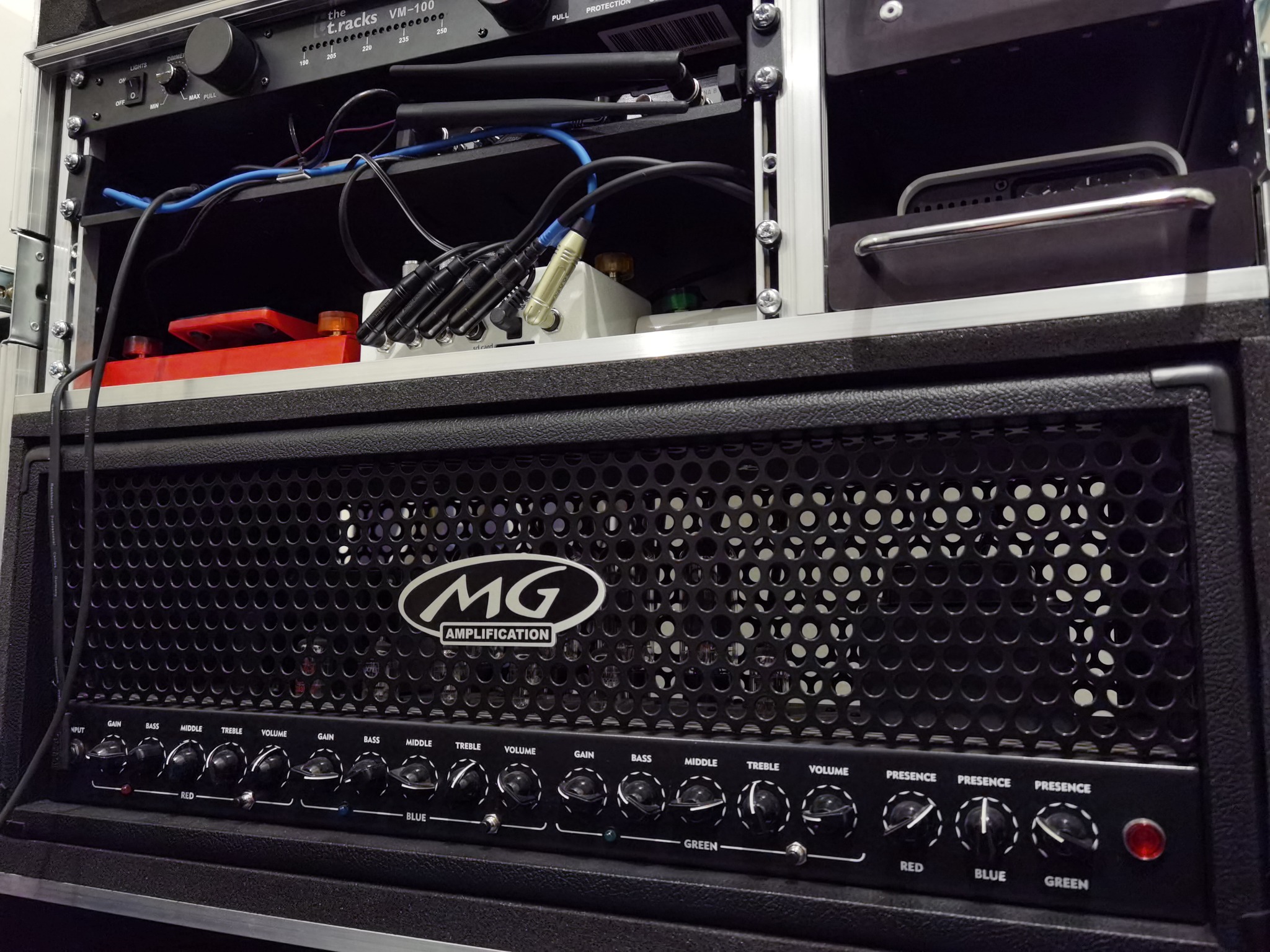 Czarny wzmacniacz gitarowy MG Amplification z widocznymi regulatorami gain, bass, middle, treble, volume, presence, umieszczony w racku z kablami i innymi urządzeniami.