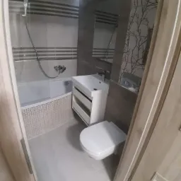 Nowoczesna łazienka z wanną obudowaną mozaiką, umywalką z szafką i podwieszaną toaletą, wykończona płytkami imitującymi drewno i szarymi płytkami z poziomymi paskami oraz motywem drzew na ścianie...