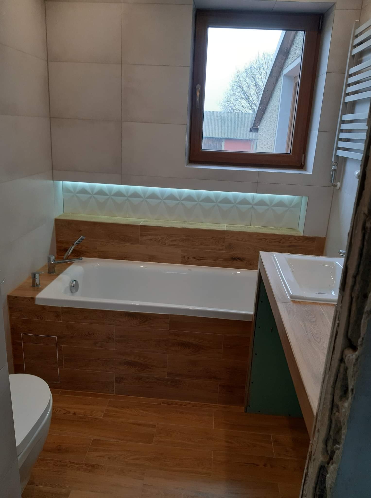 Wykończona łazienka z wanną obudowaną płytkami imitującymi drewno, podświetlaną wnęką nad wanną, białymi płytkami na ścianach i drewnianą podłogą; widok na okno z widokiem na budynki.