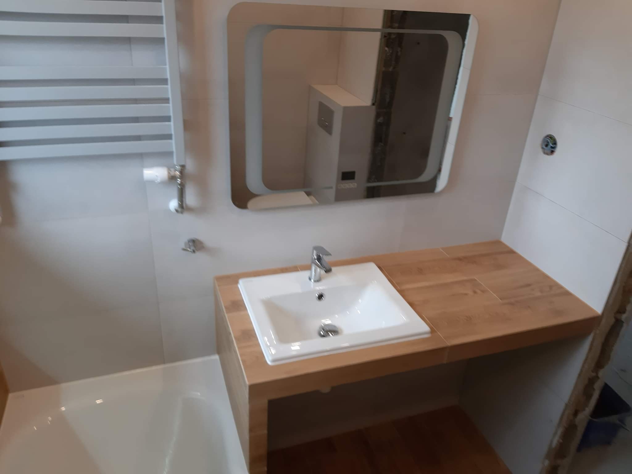 Nowoczesna łazienka po remoncie: biała umywalka nablatowa na drewnianym blacie, podświetlane lustro z zaokrąglonymi rogami, grzejnik drabinkowy i fragment wanny.