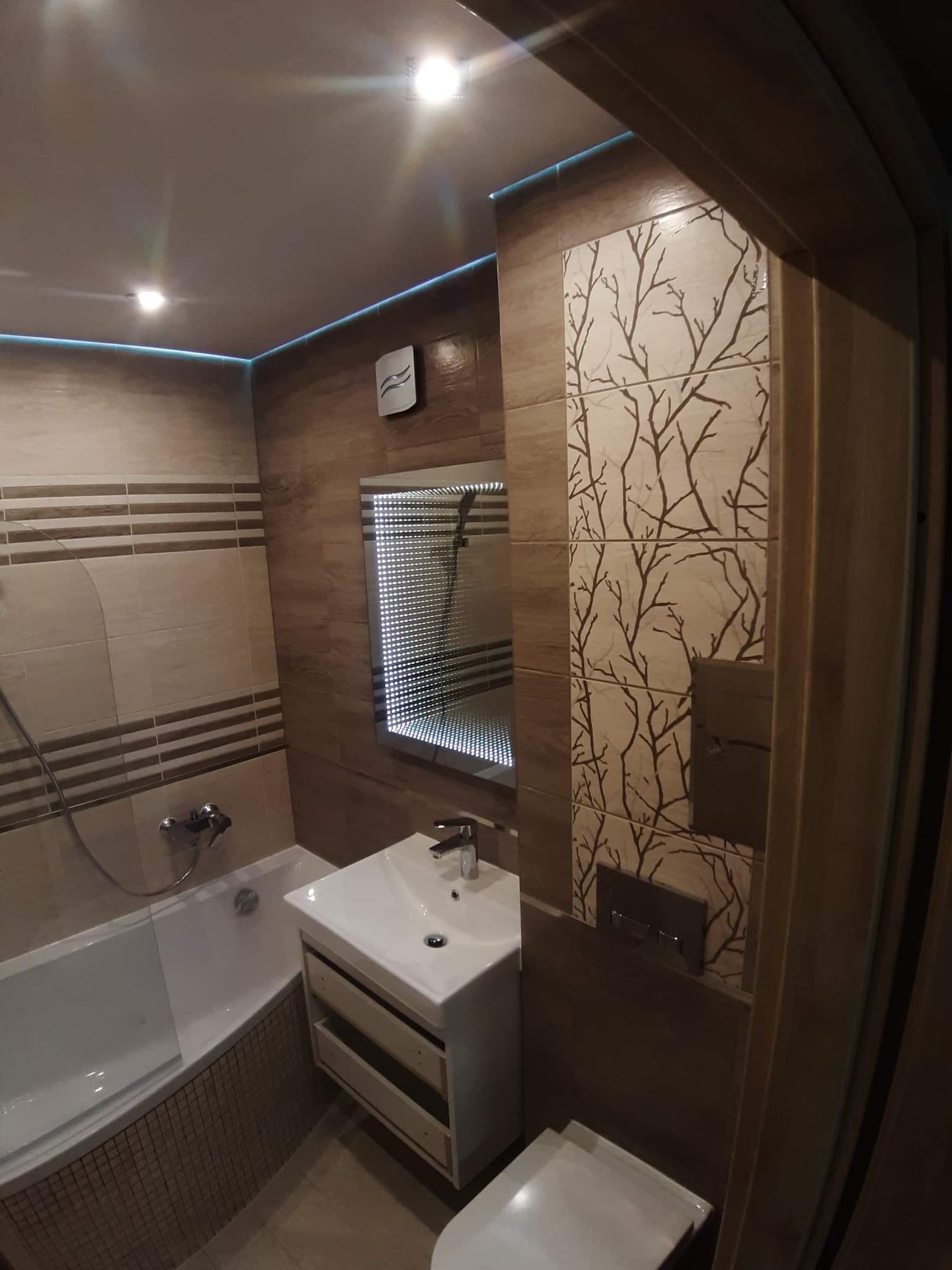 Wykończona łazienka w brązach z podświetleniem LED na suficie, dekoracyjnymi płytkami z motywem drzewa, lustrem z efektem głębi, białą umywalką z szafką i wanną z mozaiką.