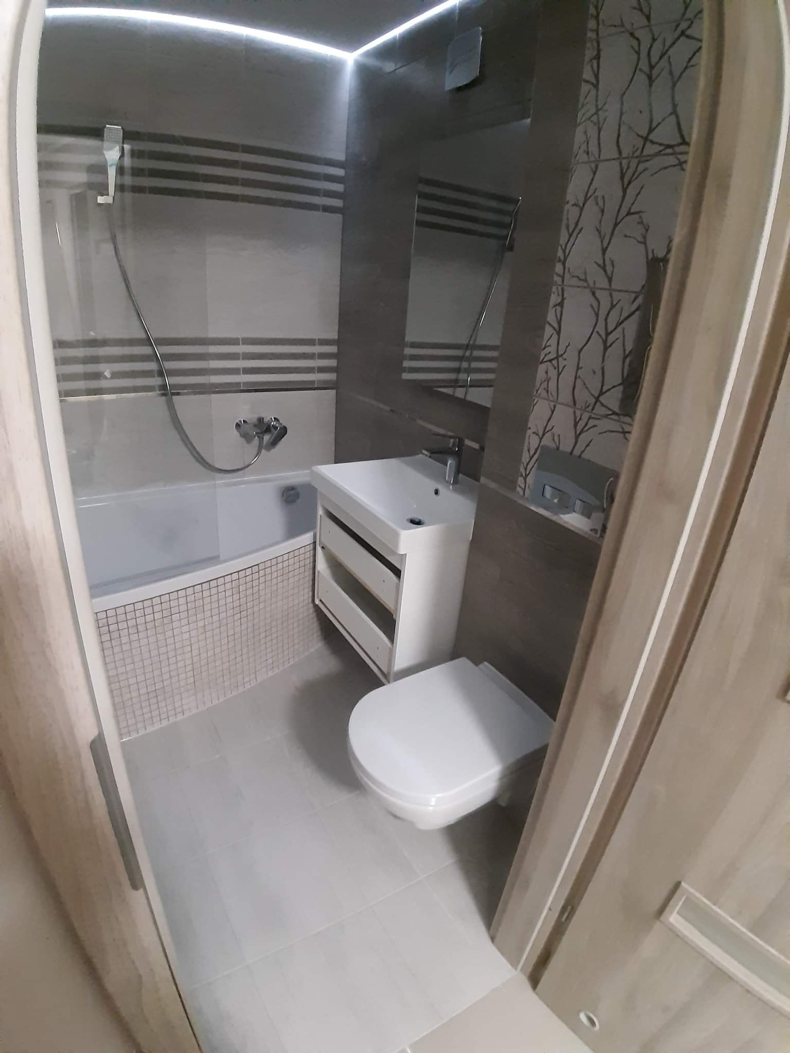 Nowoczesna łazienka z wanną obudowaną mozaiką, umywalką z szafką i podwieszaną toaletą, wykończona płytkami imitującymi drewno i szarymi płytkami z poziomymi paskami oraz motywem drzew na ścianie...