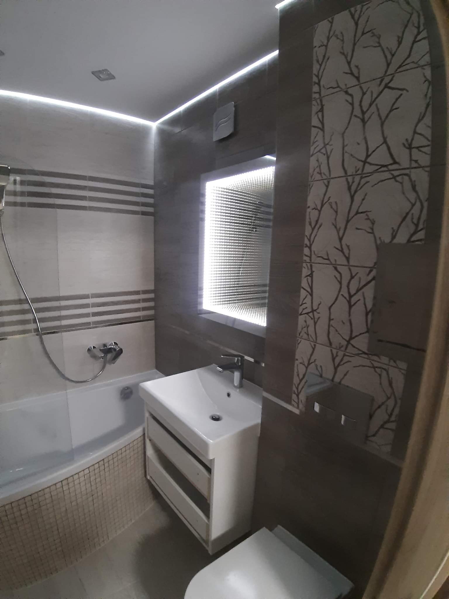 Nowoczesna łazienka z podświetlanym lustrem, płytkami imitującymi drzewo na ścianie, umywalką z szafką i wanną obudowaną mozaiką.