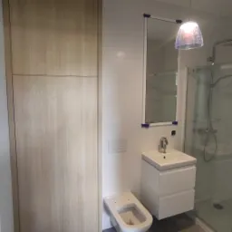 Nowoczesna łazienka z szarą marmurową podłogą, wiszącą toaletą, białą szafką z umywalką, lustrem i kabiną prysznicową, oświetlona lampą wiszącą z przezroczystym kloszem.