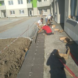 PROTOCLUSTER SP. Z O.O. - Trzech mężczyzn układa ciemnoszare płytki chodnikowe na zewnątrz budynku mieszkalnego; widoczne kartony z płytkami, poziomice i ziemia.