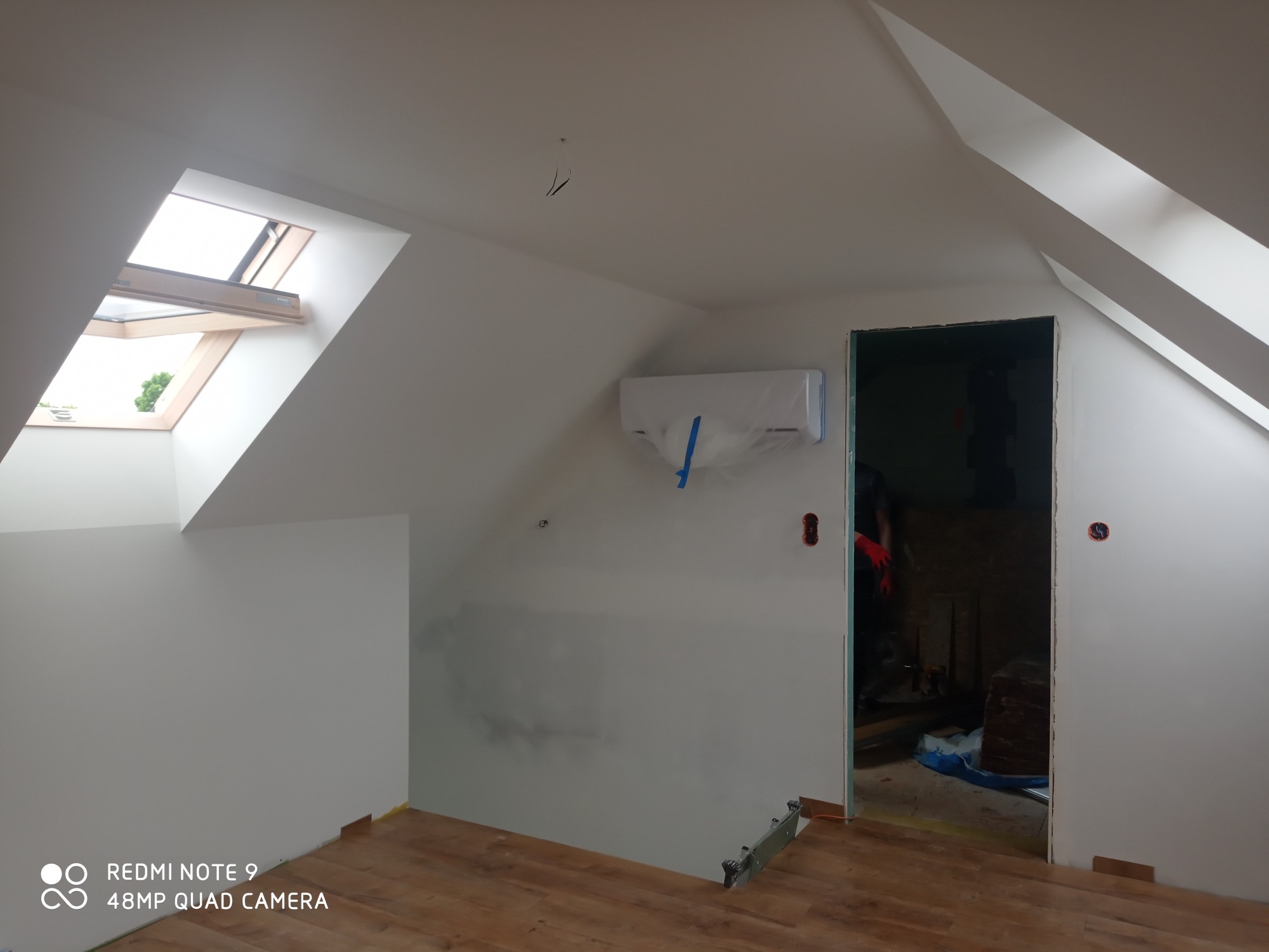 Poddasze w trakcie prac wykończeniowych: ściany po nałożeniu gładzi, częściowo pomalowane na biało, okno dachowe, zamontowany klimatyzator zabezpieczony folią, podłoga wyłożona panelami, widoczne...