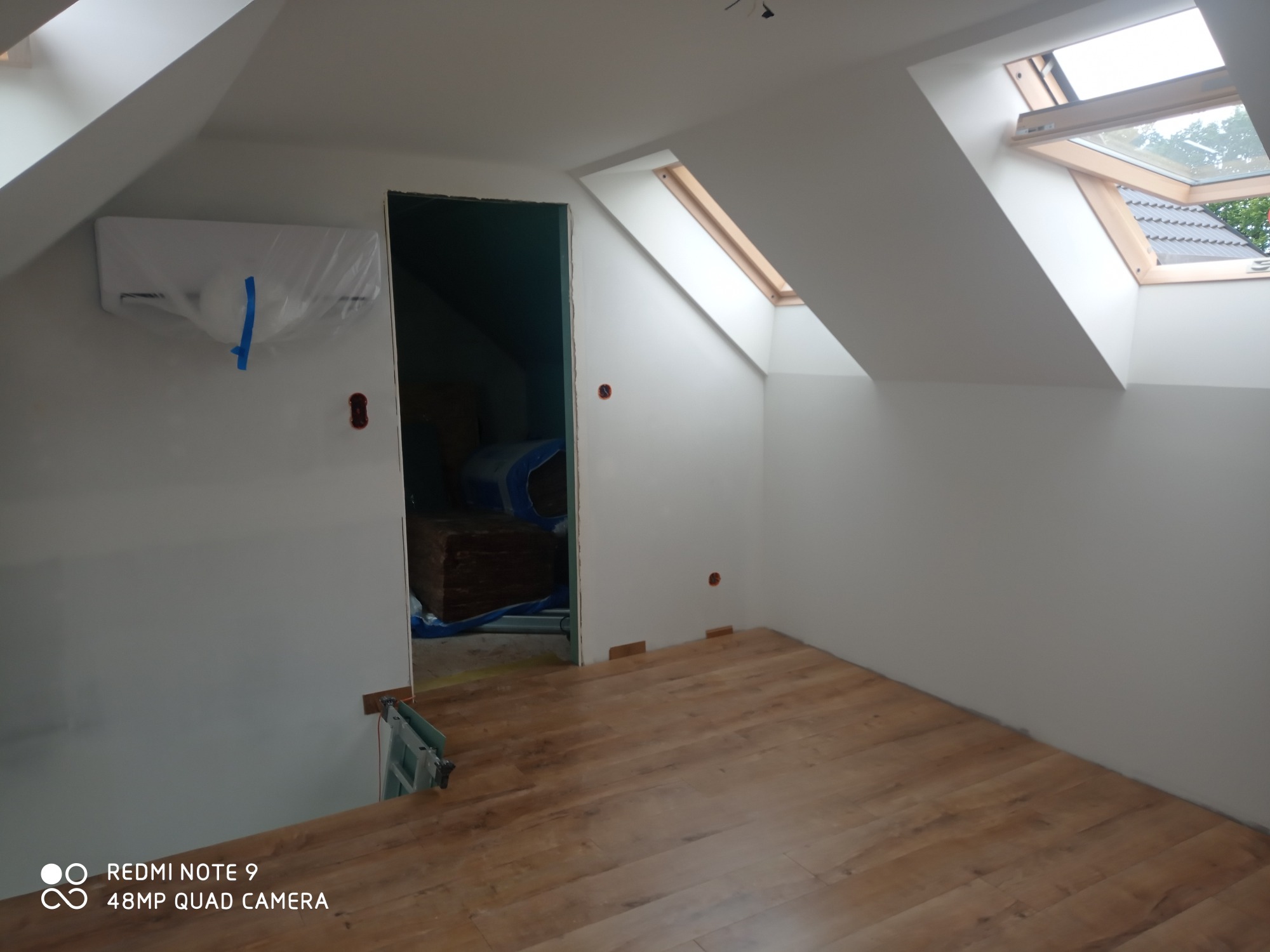 Poddasze po remoncie: świeżo pomalowane ściany na biało, drewnopodobne panele na podłodze, okna dachowe z drewnianą ramą, klimatyzator zabezpieczony folią. Widoczne przygotowane panele do położenia...