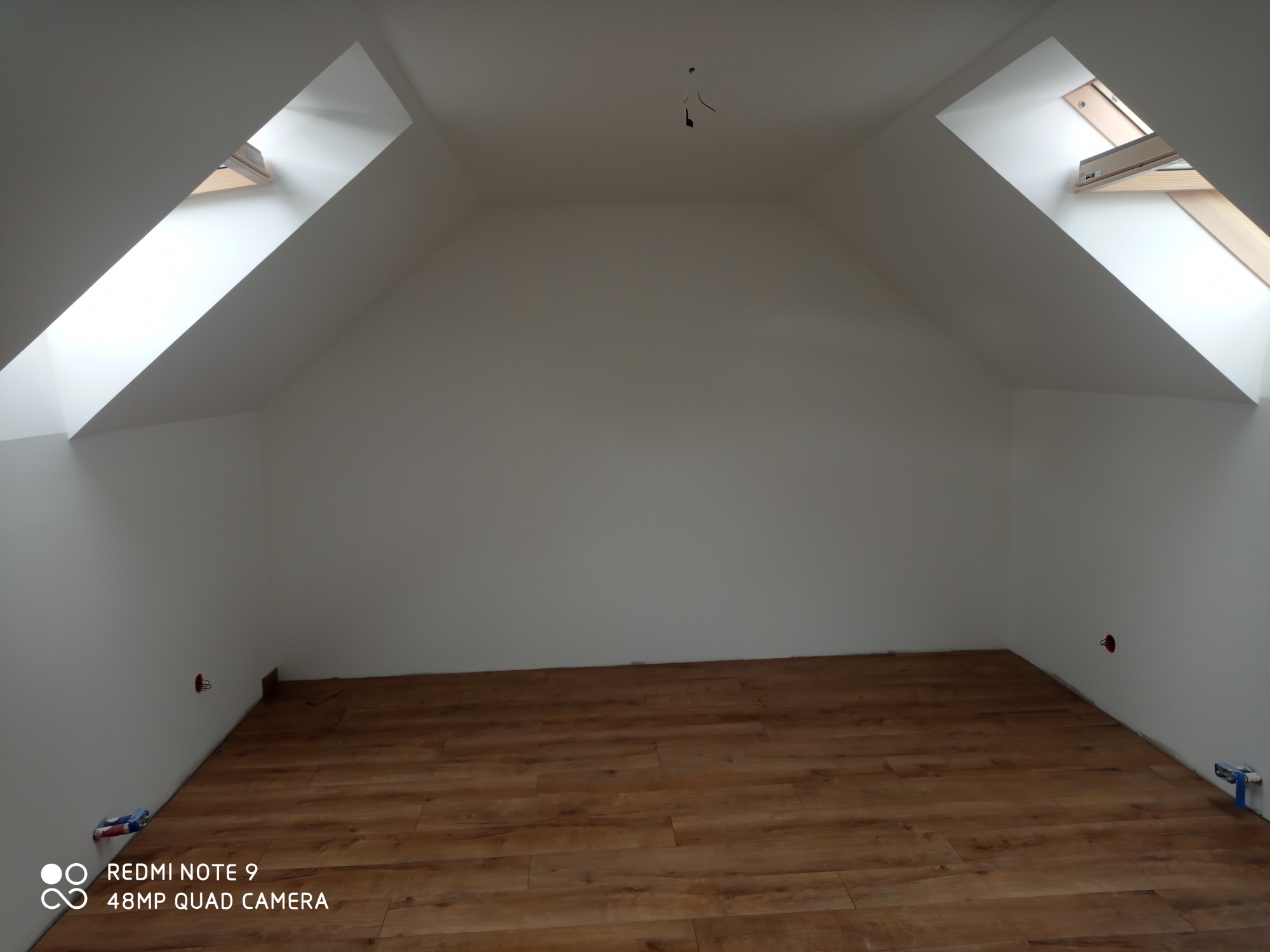 Poddasze po remoncie: ułożone panele podłogowe w odcieniach brązu, białe ściany i dwa okna dachowe w skosach dachu, widoczne puszki elektryczne i wystające kable.