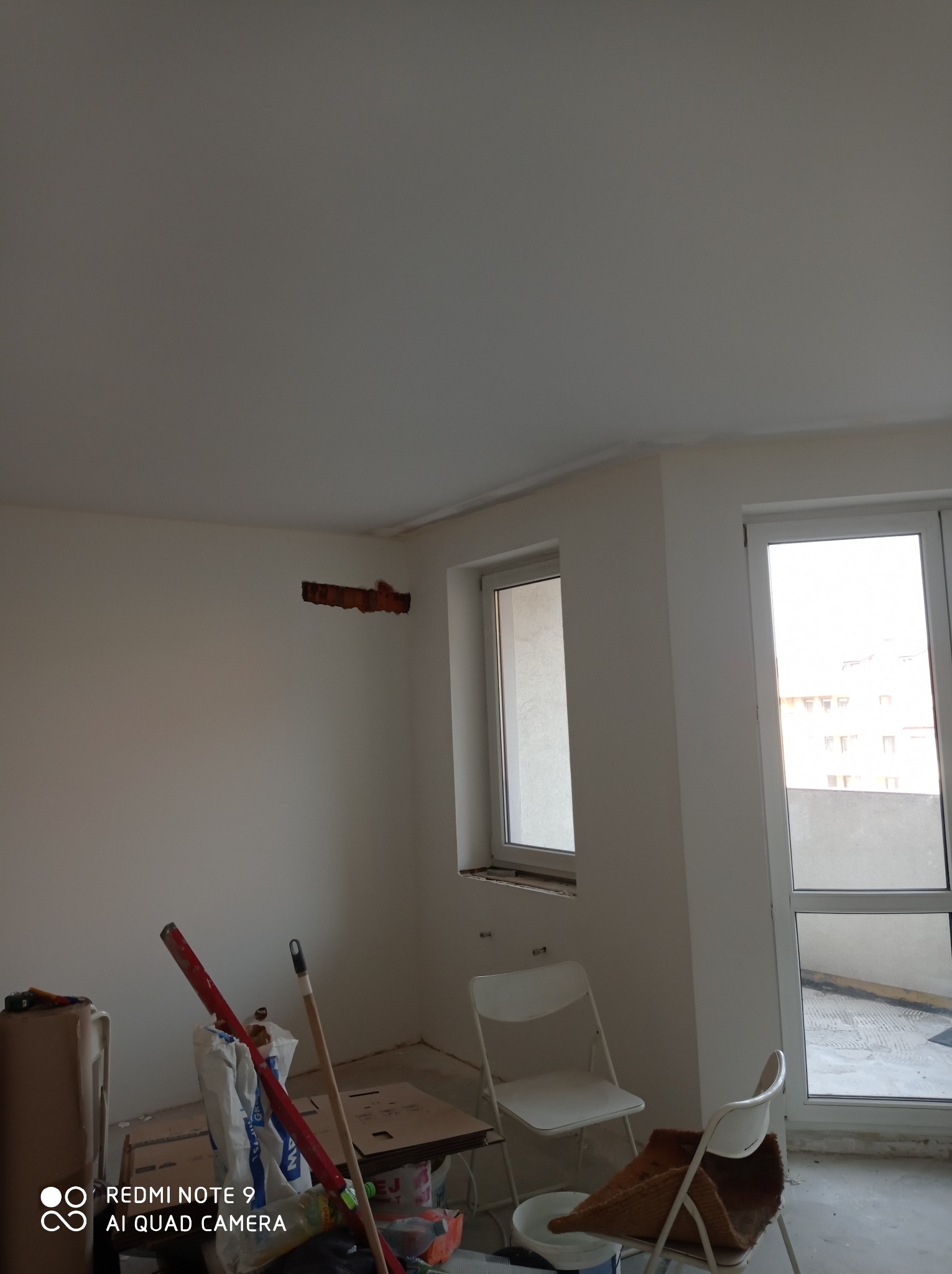 Wnętrze w trakcie prac przygotowawczych: ściana z wyciętym otworem, widoczne narzędzia i materiały budowlane, dwa białe krzesła składane, okno i drzwi balkonowe.
