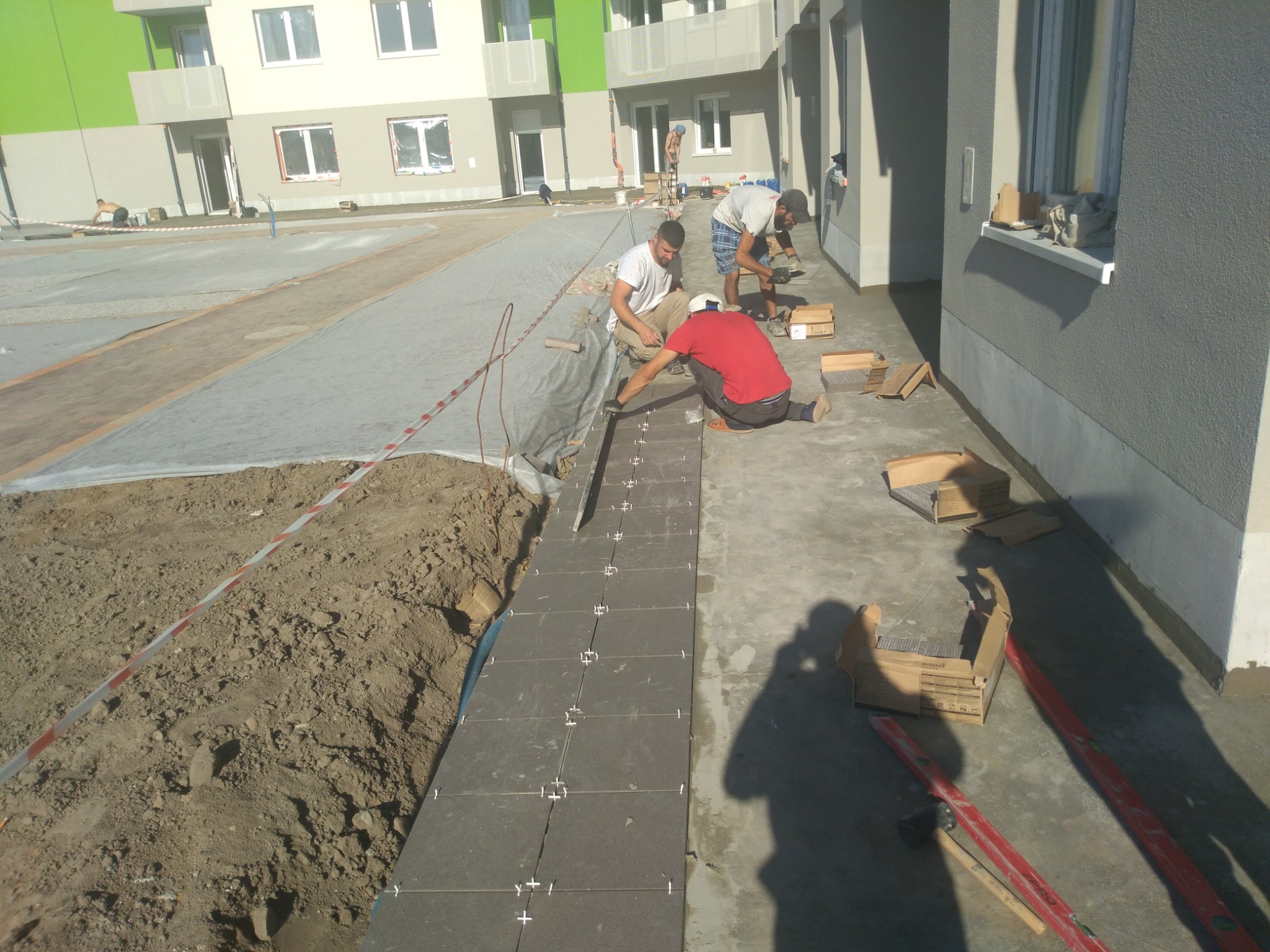 Trzech mężczyzn układa ciemnoszare płytki chodnikowe na zewnątrz budynku mieszkalnego; widoczne kartony z płytkami, poziomice i ziemia.