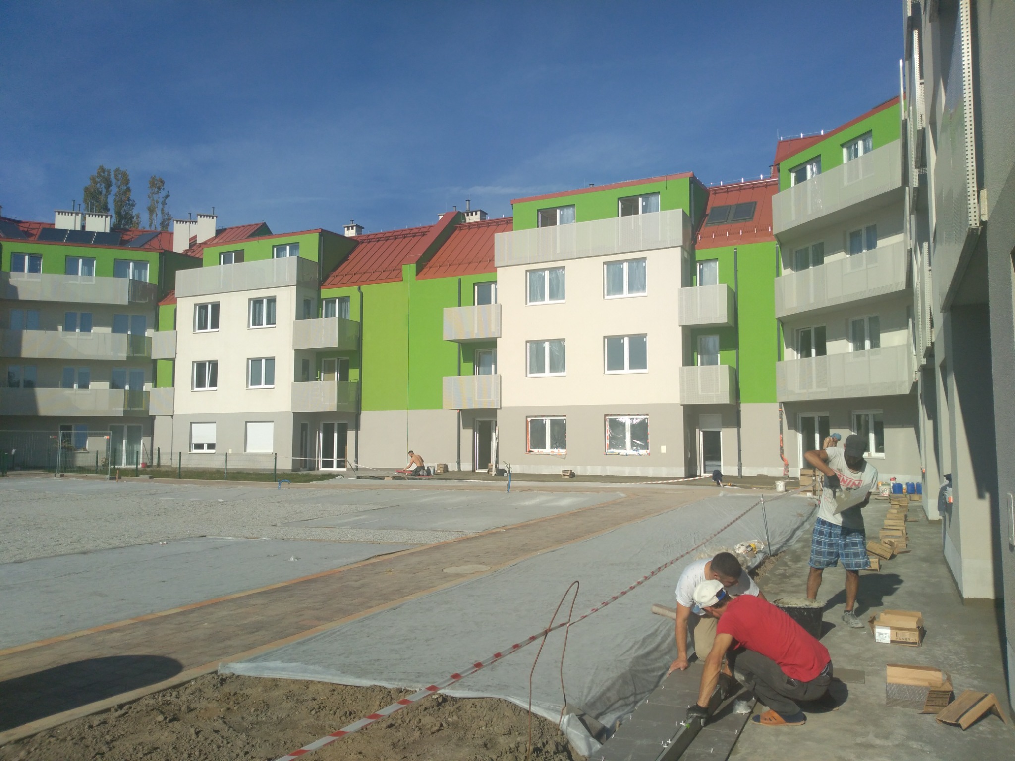 Układanie kostki brukowej na przygotowanym podłożu przed nowym budynkiem mieszkalnym z zielono-białą elewacją i czerwonym dachem; widoczni pracownicy w trakcie pracy.