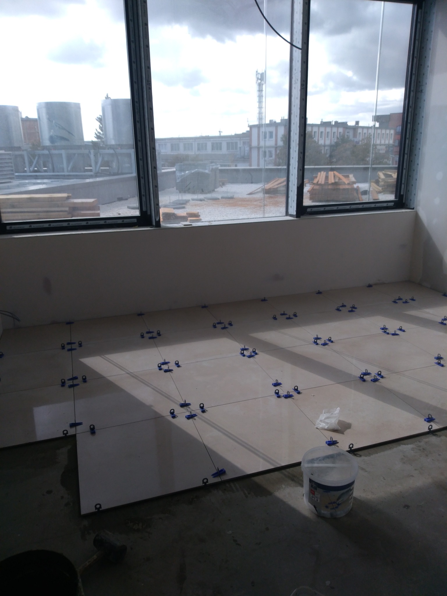 Układanie płytek podłogowych w pomieszczeniu, widoczne krzyżyki dystansowe, wiadro z klejem, oraz widok z okna na budynki i zbiorniki przemysłowe