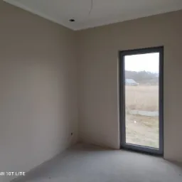 Pomalowane na beżowo wnętrze pokoju z dwoma szarymi oknami, widok na zewnątrz na pole i budynek gospodarczy, surowy beton na podłodze, brak mebli.