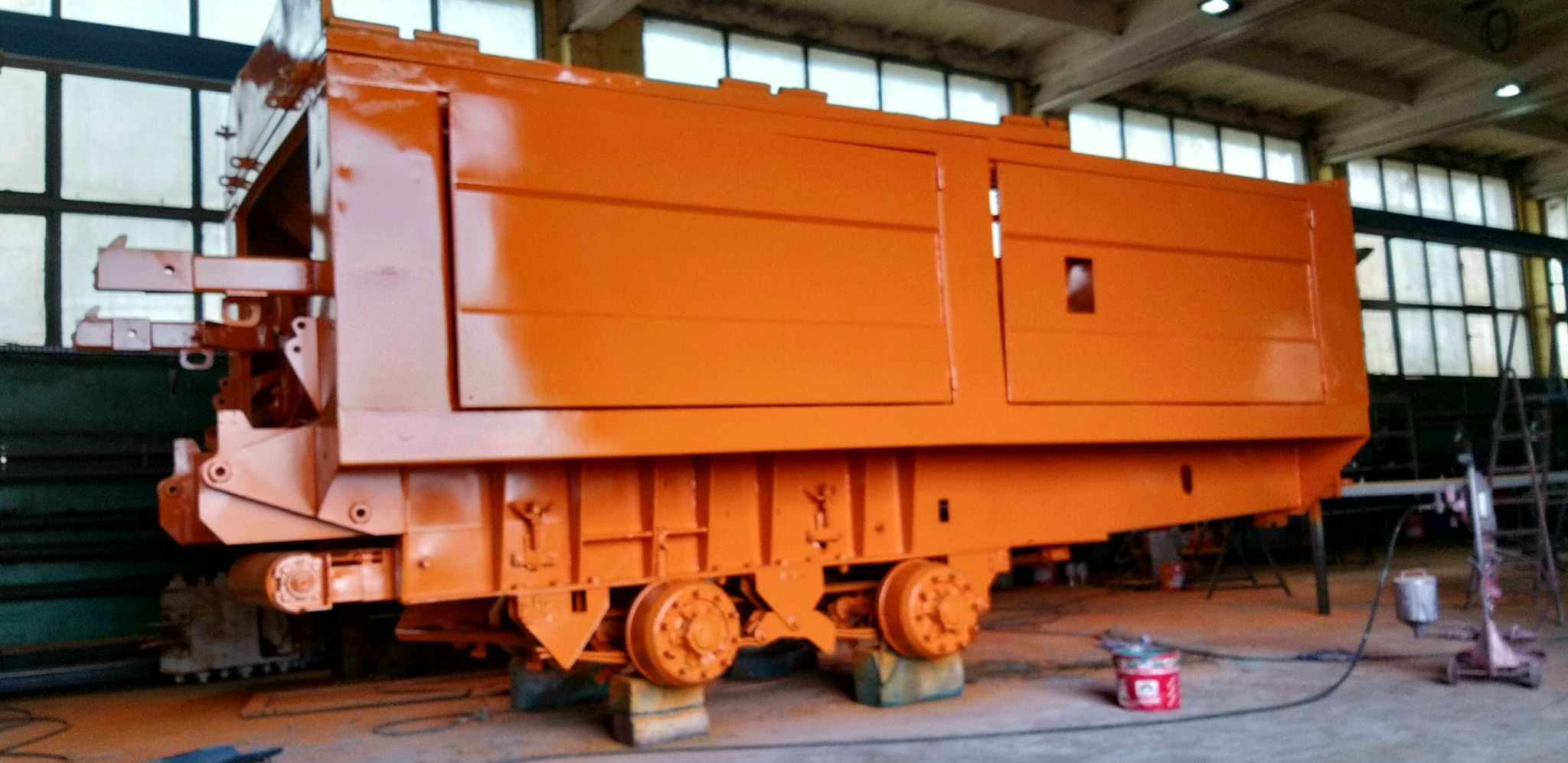 Pomarańczowy wagon kolejowy na betonowych blokach w hali produkcyjnej, świeżo po malowaniu natryskowym, widoczne ślady farby w powietrzu i puszki z farbą.