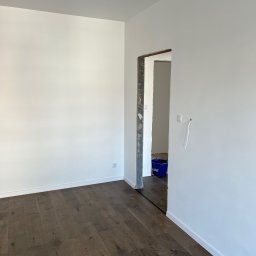 Fams bud - Pusty pokój z nową, ciemną drewnianą podłogą, świeżo pomalowanymi na biało ścianami i surowym otworem drzwiowym, widoczny niebieski kubeł w drugim pomieszczeniu.
