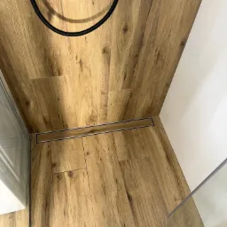 Parkiet w łazience z odpływem liniowym, kabina prysznicowa ze szklaną ścianką, białe płytki na ścianie. Nowoczesne wykończenie wnętrza łazienki.