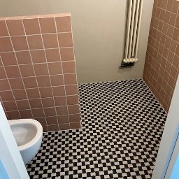 Fams bud - Wykończona łazienka z czarno-białą szachownicą na podłodze i różowymi płytkami na ścianie. Grzejnik żeberkowy na ścianie. Widok zza framugi drzwi.