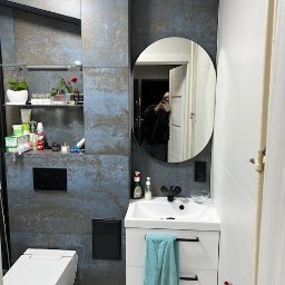 Fams bud - Nowoczesna łazienka z płytkami imitującymi rdzę, owalnym lustrem i białą szafką umywalkową z turkusowym ręcznikiem. Widoczna toaleta i półki z kosmetykami.