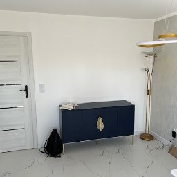 Fams bud - Nowoczesne wnętrze: granatowa komoda z złotymi uchwytami, designerska lampa podłogowa i drzwi z poziomymi przeszkleniami. Minimalistyczny styl, jasne ściany i marmurowa podłoga.