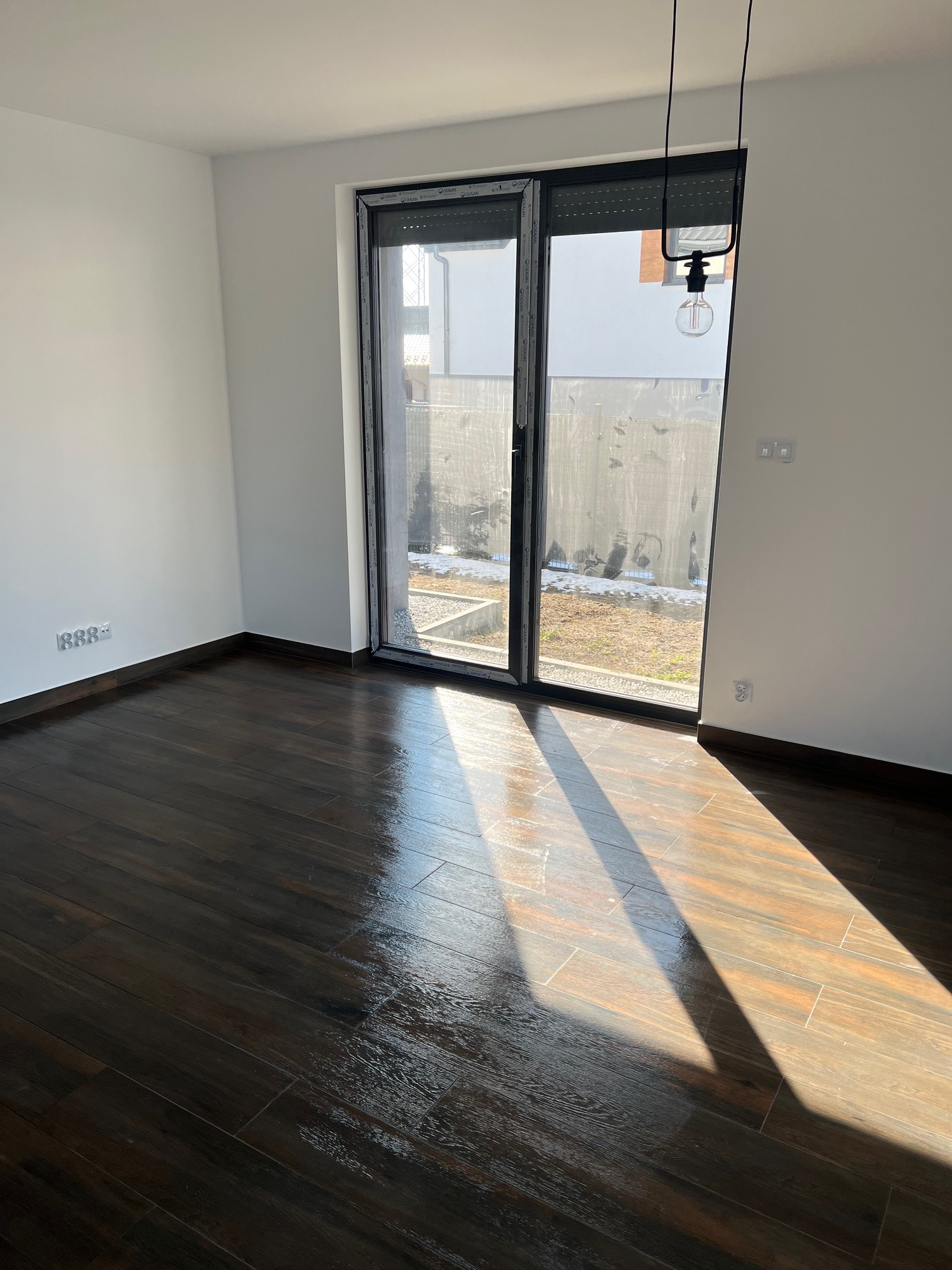 Pusty pokój z ciemną, błyszczącą podłogą z płytek imitujących drewno, duże okno przesuwne z widokiem na ogród i minimalistyczną lampą wiszącą z żarówką Edisona.