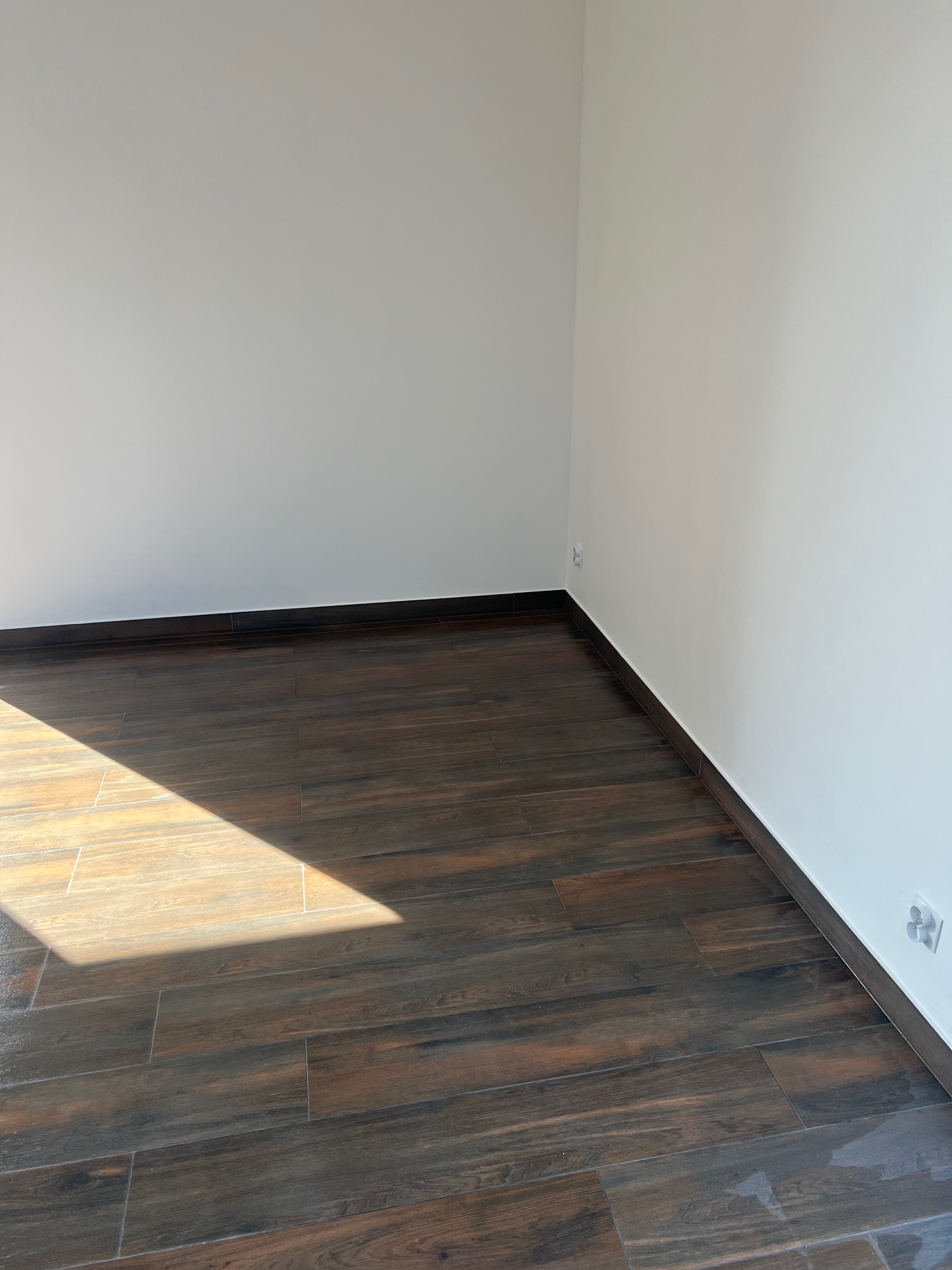 Pusty pokój z nową, ciemną podłogą imitującą drewno, białe ściany i widoczne gniazdko elektryczne, oświetlony naturalnym światłem wpadającym przez okno.