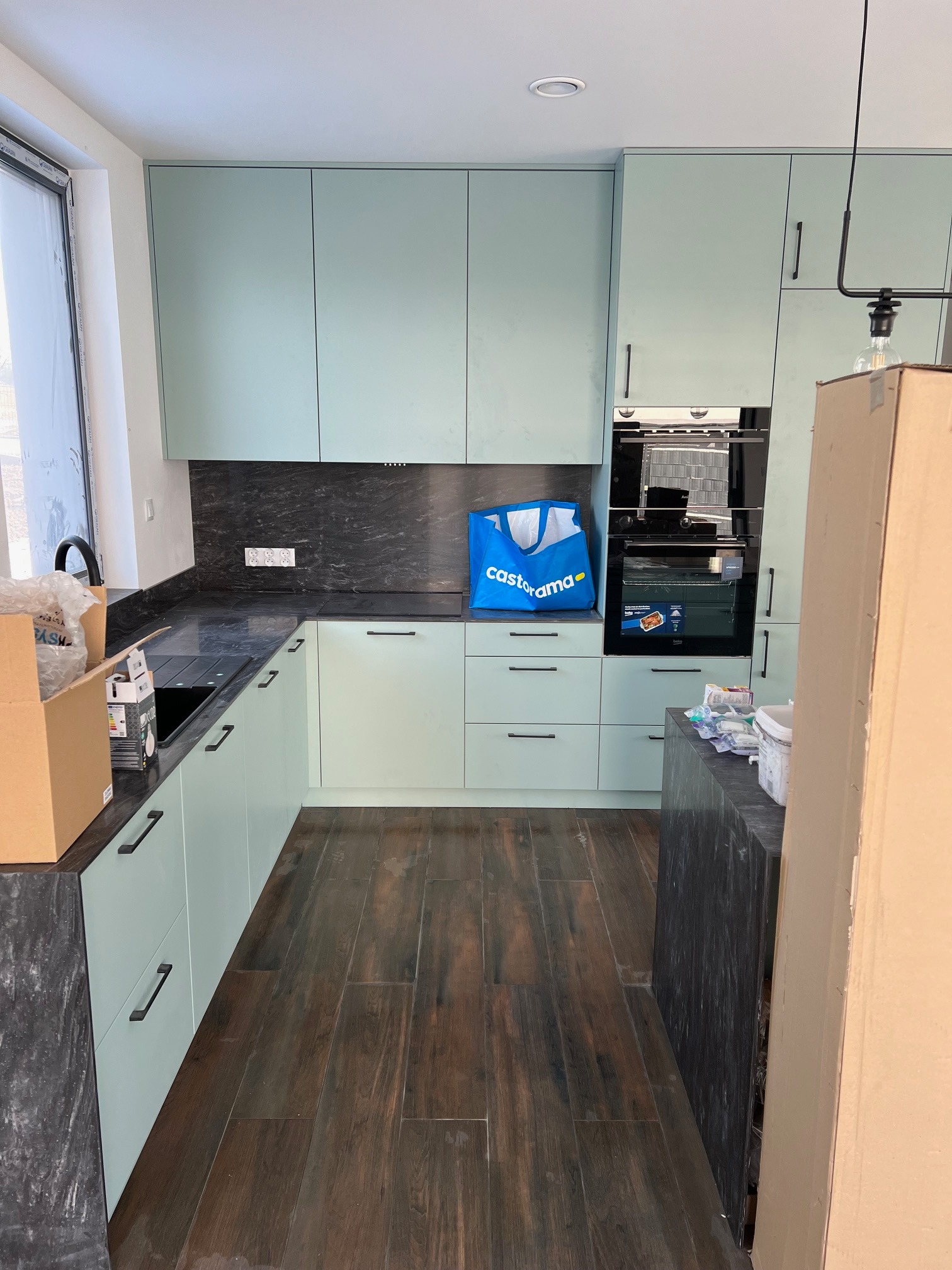 Nowoczesna kuchnia z jasnymi, miętowo-zielonymi szafkami, czarnym blatem i drewnopodobną podłogą w trakcie montażu. Widoczna torba Castorama na blacie, kartony i materiały wykończeniowe sugerują...