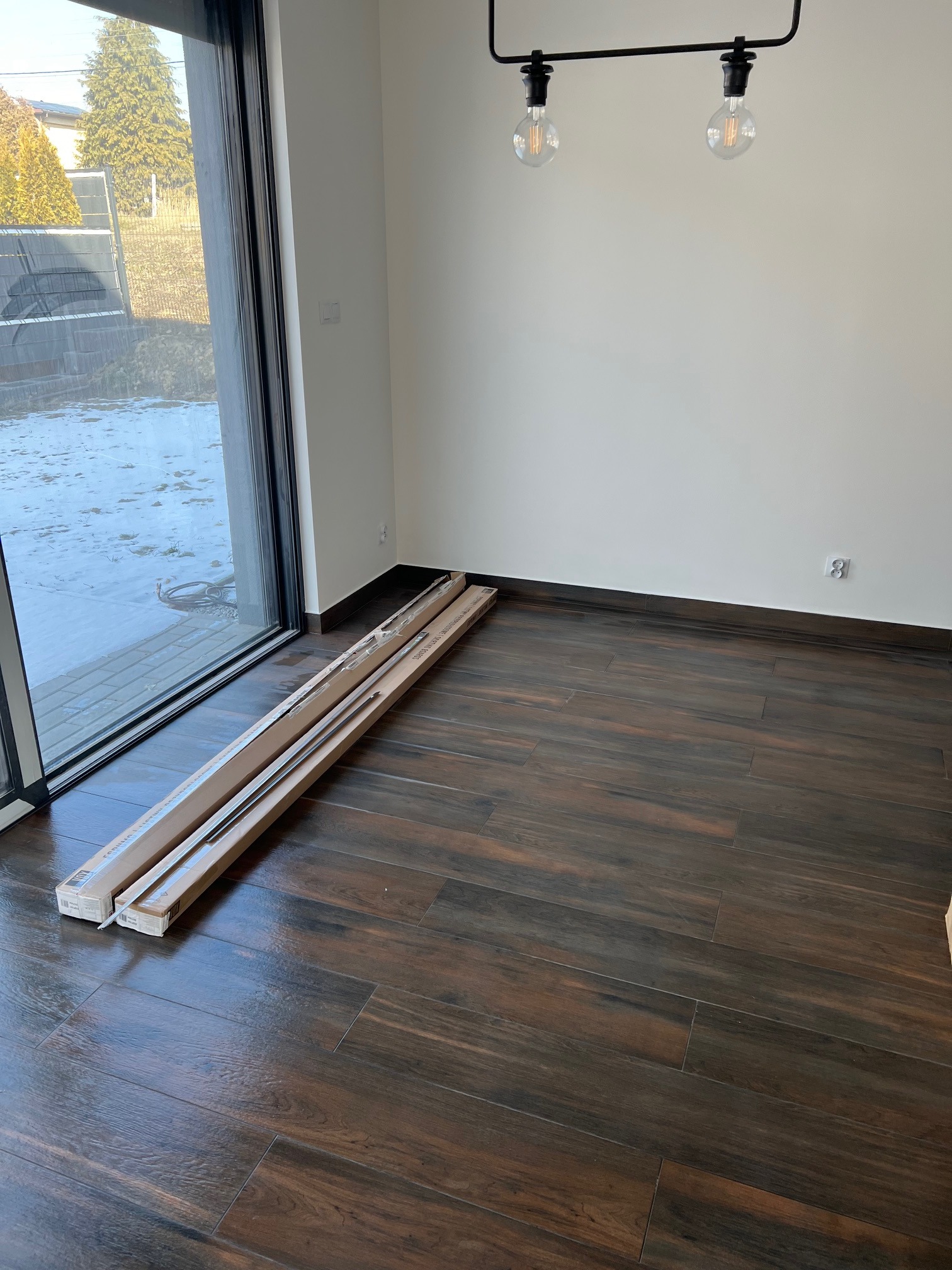 Nowe, zapakowane listwy przypodłogowe w kartonach leżące na podłodze z ciemnego drewna w nowym pokoju z dużym oknem przesuwnym i minimalistyczną lampą sufitową.