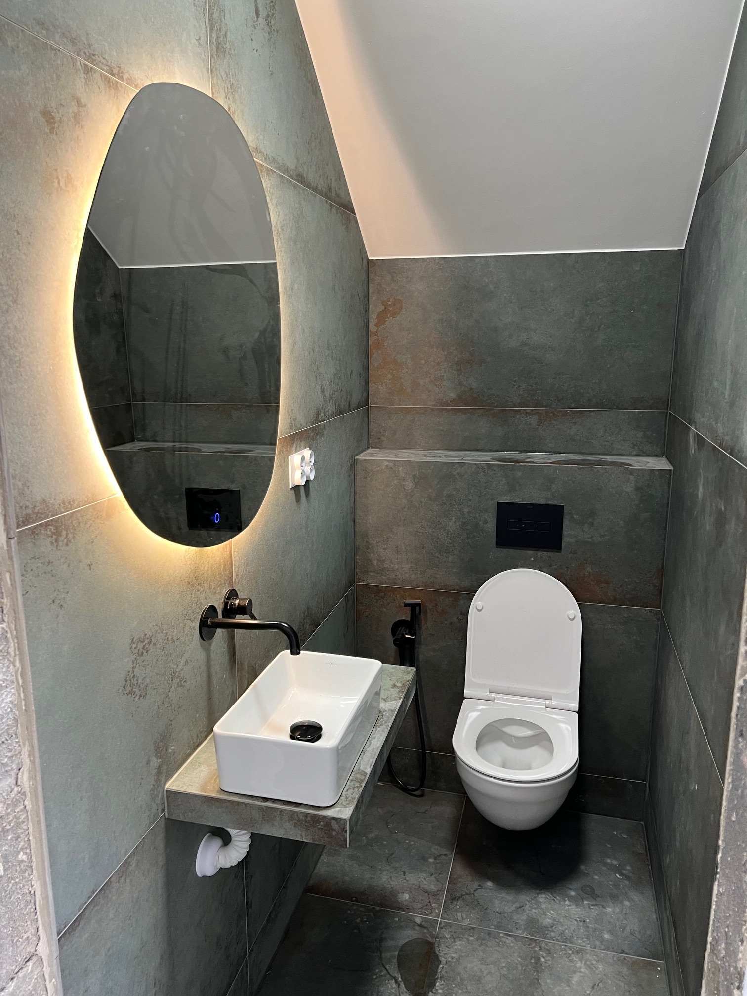 Nowoczesna łazienka z płytkami imitującymi kamień, podświetlanym owalnym lustrem, białą umywalką nablatową na kamiennym blacie i wiszącą toaletą.