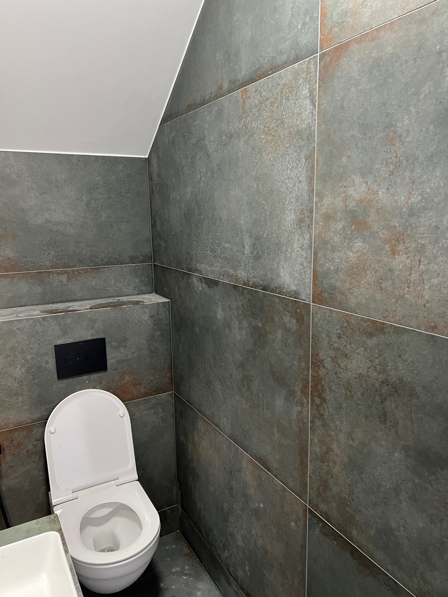 Nowoczesna toaleta z szarymi płytkami imitującymi rdzę, biała ceramika sanitarna i czarny przycisk spłuczki pod skosem sufitu.