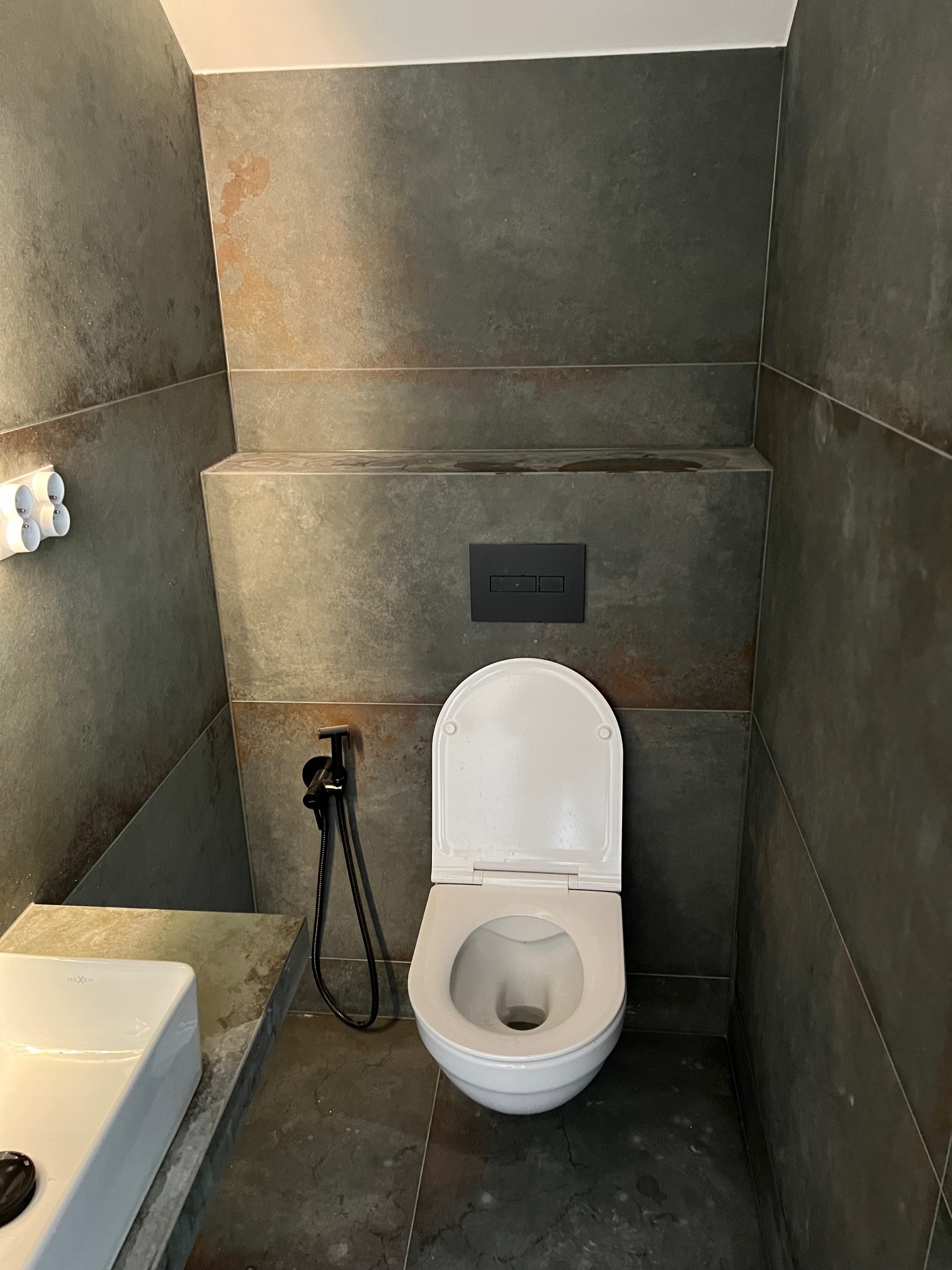 Nowoczesna toaleta z miską WC z otwartą klapą, bidetką i umywalką w minimalistycznym wnętrzu wykończonym grafitowymi płytkami imitującymi beton.