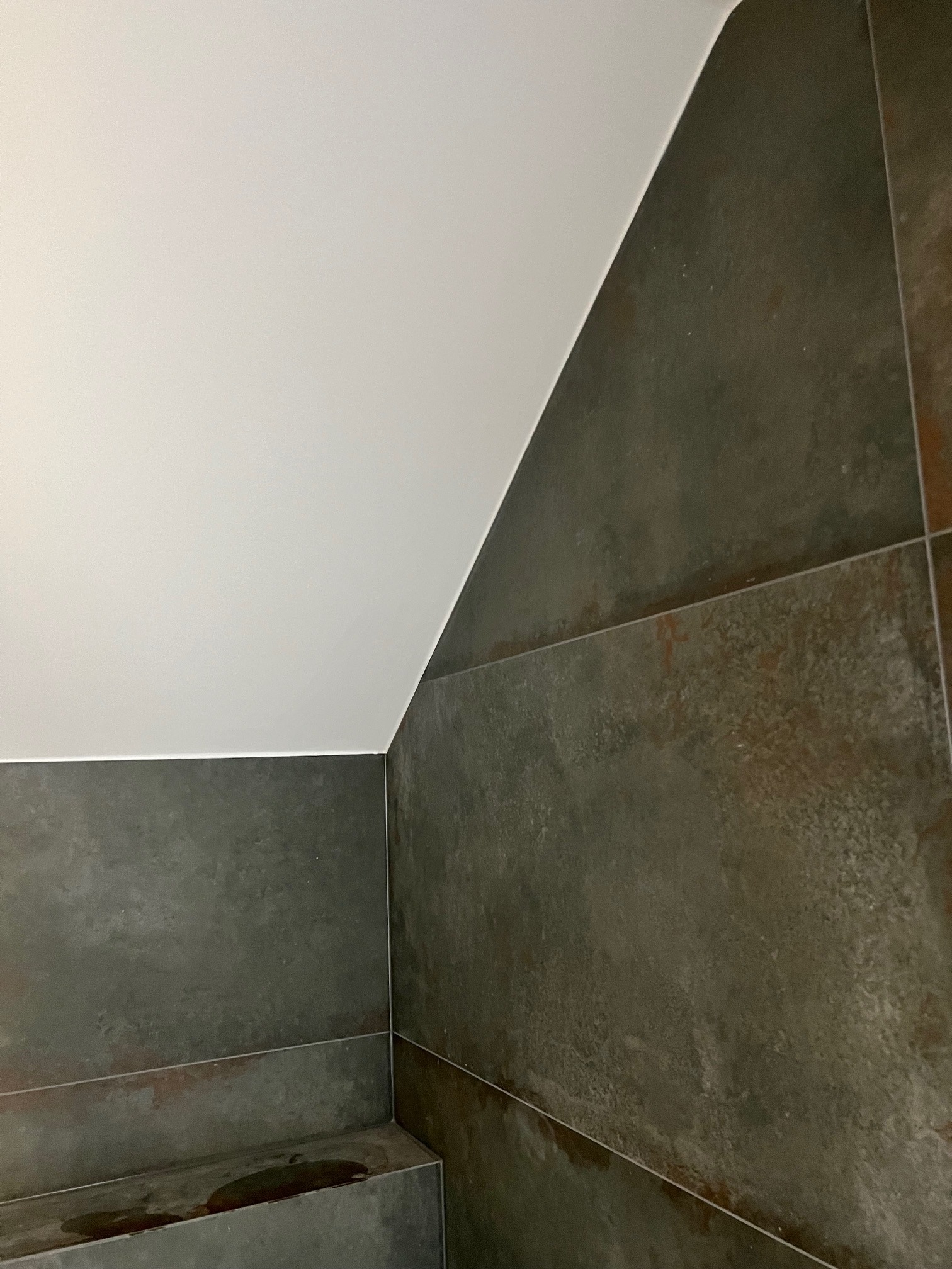 Szczegół wykończenia łazienki z ciemnymi płytkami imitującymi rdzę, biały sufit ze skosem, widoczne fugi i plama wody na półce.