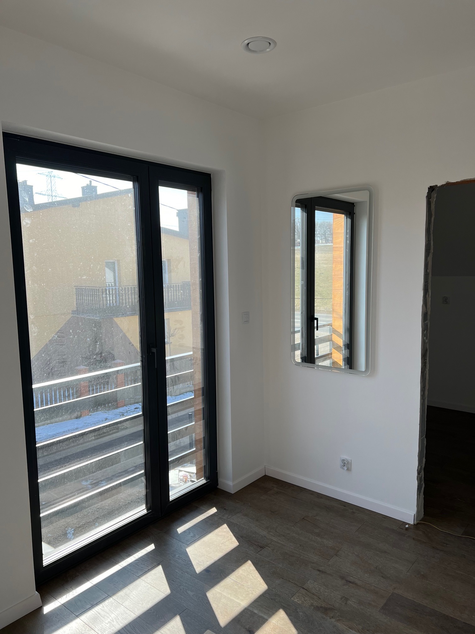 Wnętrze pokoju z dużym oknem balkonowym w czarnej ramie, odbicie okna w lustrze na ścianie, podłoga z paneli w odcieniach brązu, widoczne ślady po remoncie przy wejściu do pokoju.
