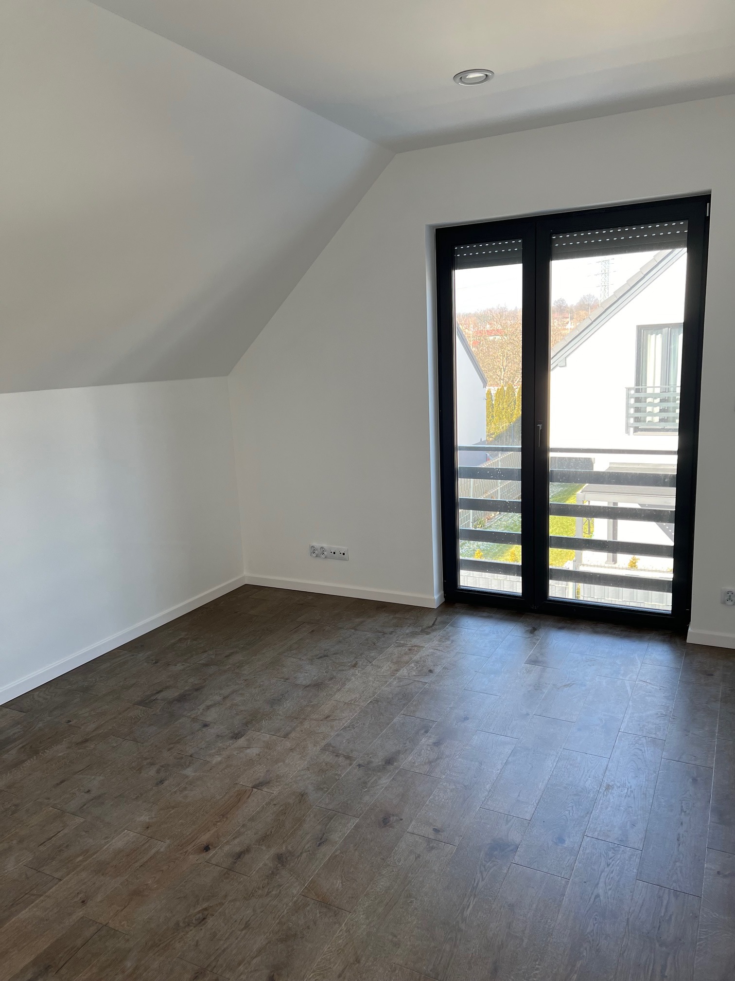 Puste poddasze z brązową podłogą imitującą drewno, białe ściany, okno balkonowe z czarnymi ramami i roletami zewnętrznymi, widok na ogród sąsiedniego domu.