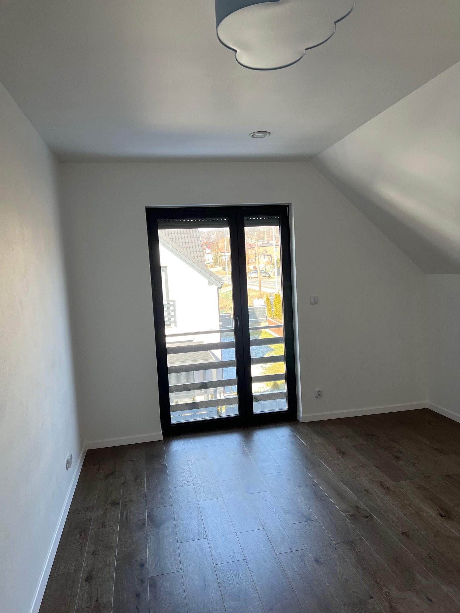 Puste poddasze z drewnianą podłogą, białymi ścianami i czarnymi drzwiami balkonowymi z widokiem na osiedle domów.