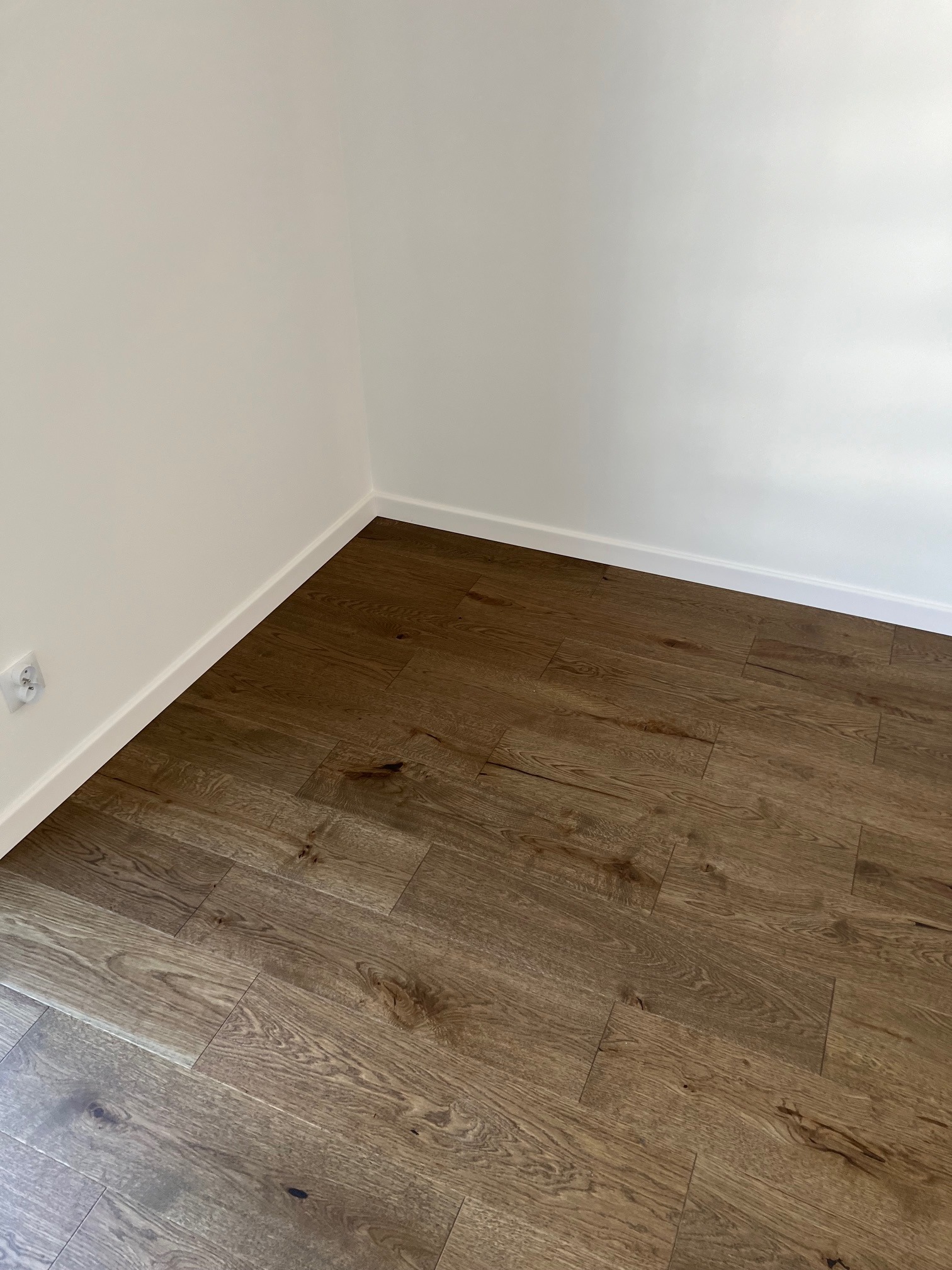 Ułożona podłoga z paneli w odcieniach brązu, białe ściany z listwami przypodłogowymi i gniazdko elektryczne po lewej stronie.