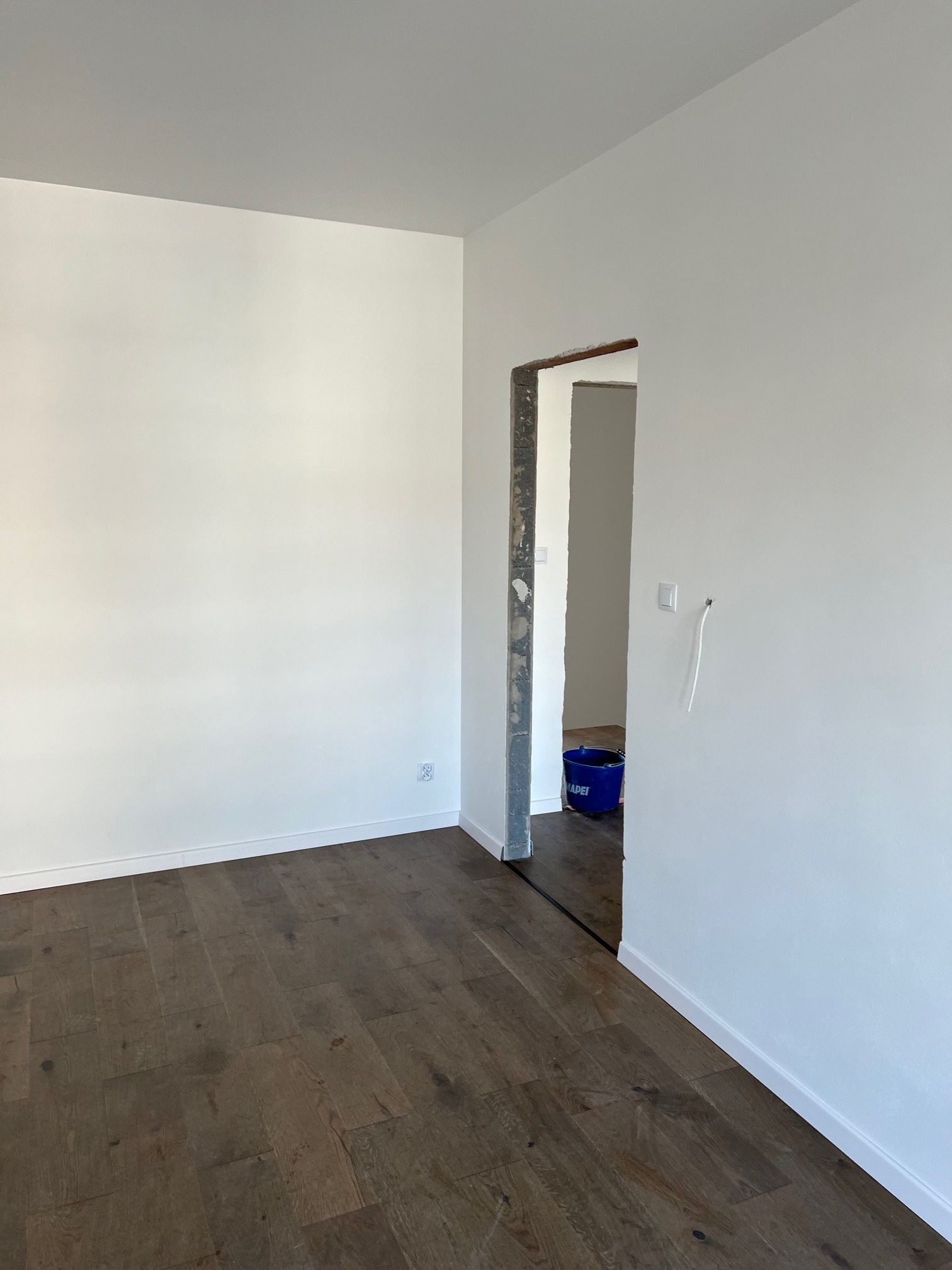 Pusty pokój z nową, ciemną drewnianą podłogą, świeżo pomalowanymi na biało ścianami i surowym otworem drzwiowym, widoczny niebieski kubeł w drugim pomieszczeniu.