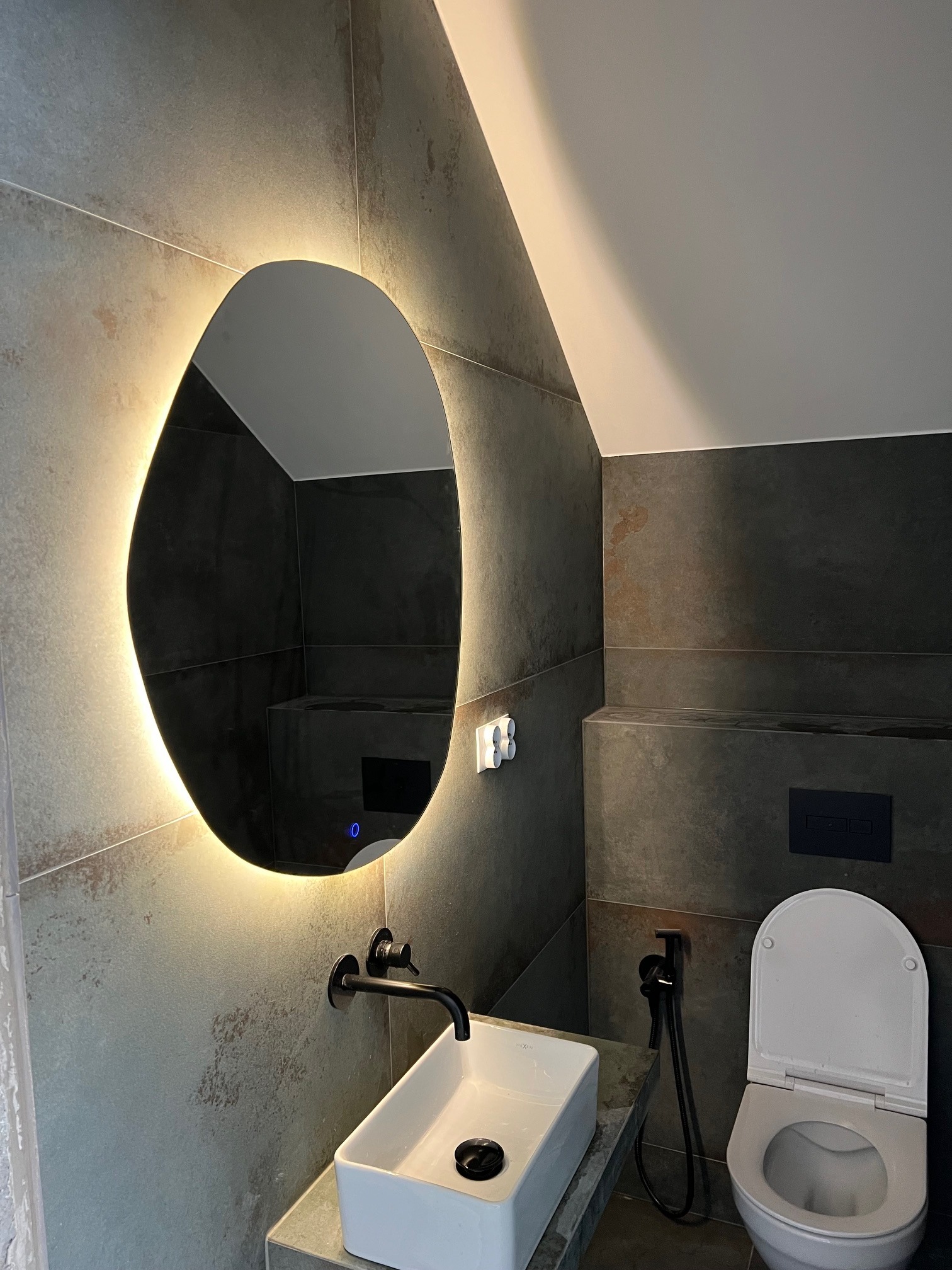 Nowoczesna łazienka z podświetlanym lustrem o nieregularnym kształcie, umywalką nablatową i toaletą podwieszaną na tle szarych płytek imitujących kamień.